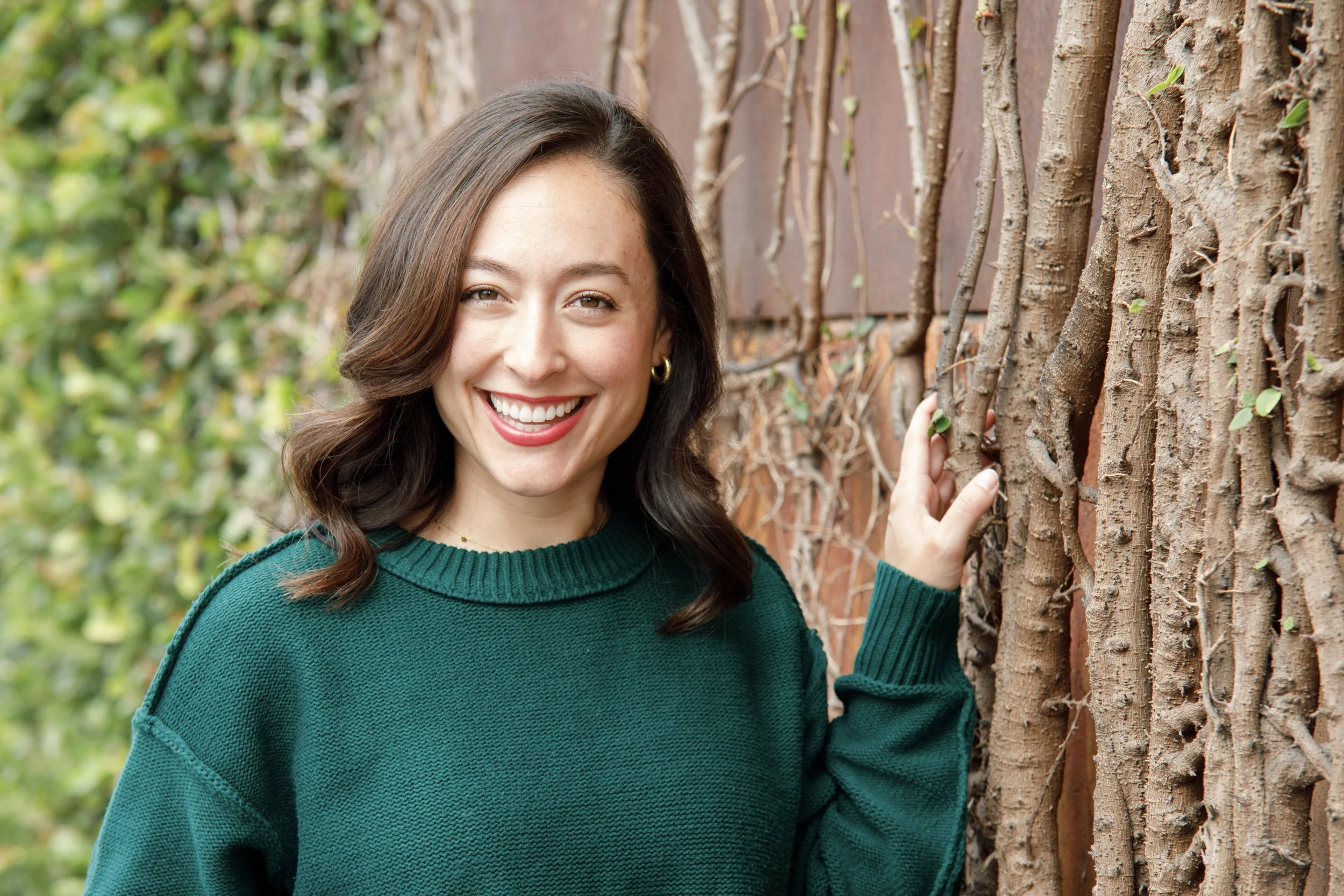 A woman with shoulder-length brown hair wearing a green sweater, smiling and touching a vine-covered brick wall outdoors.