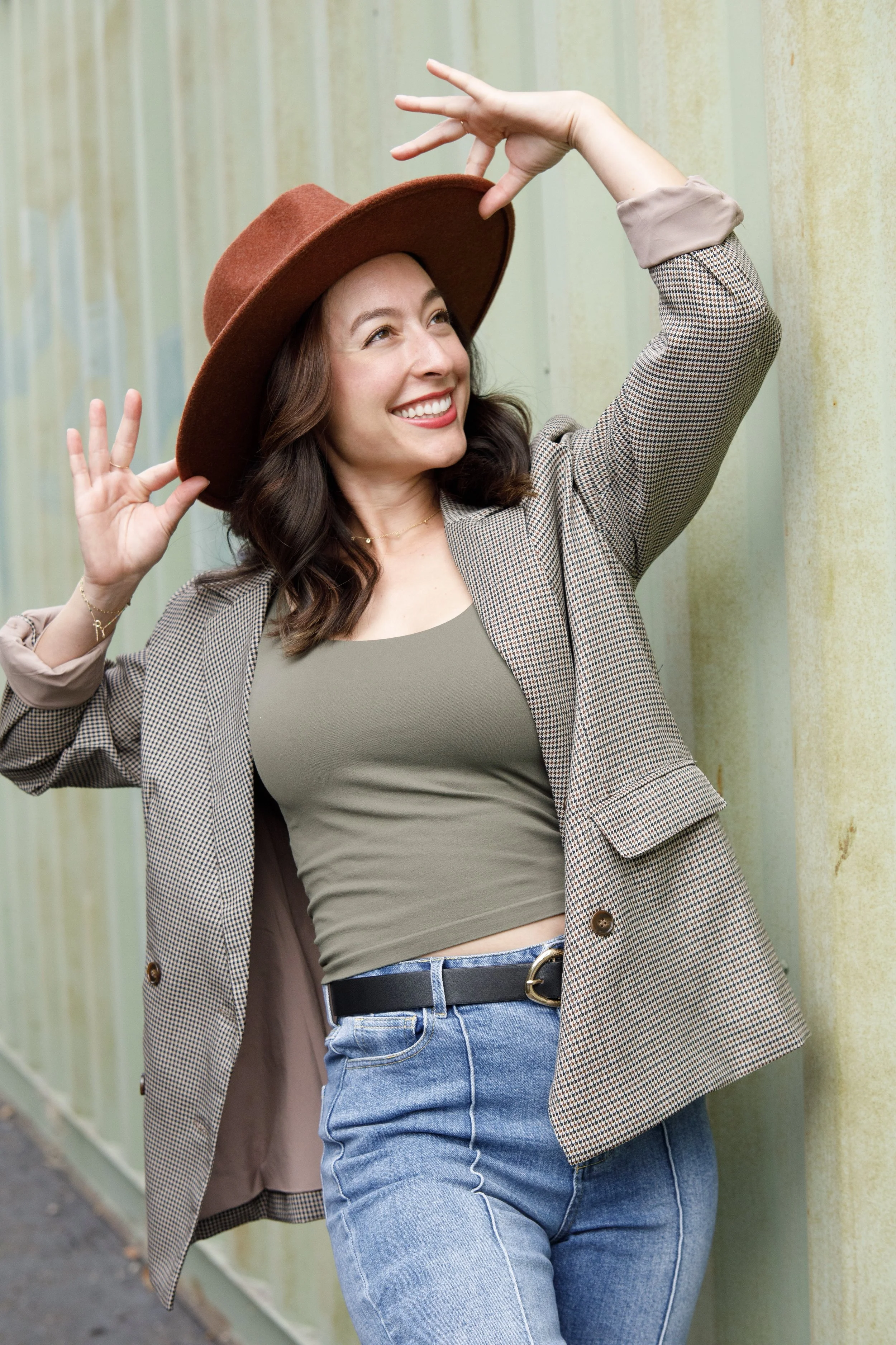 A woman with dark brown hair, smiling, wearing a brown wide-brimmed hat, a green top, a checked blazer, and blue jeans, standing outdoors against a greenish metal wall.