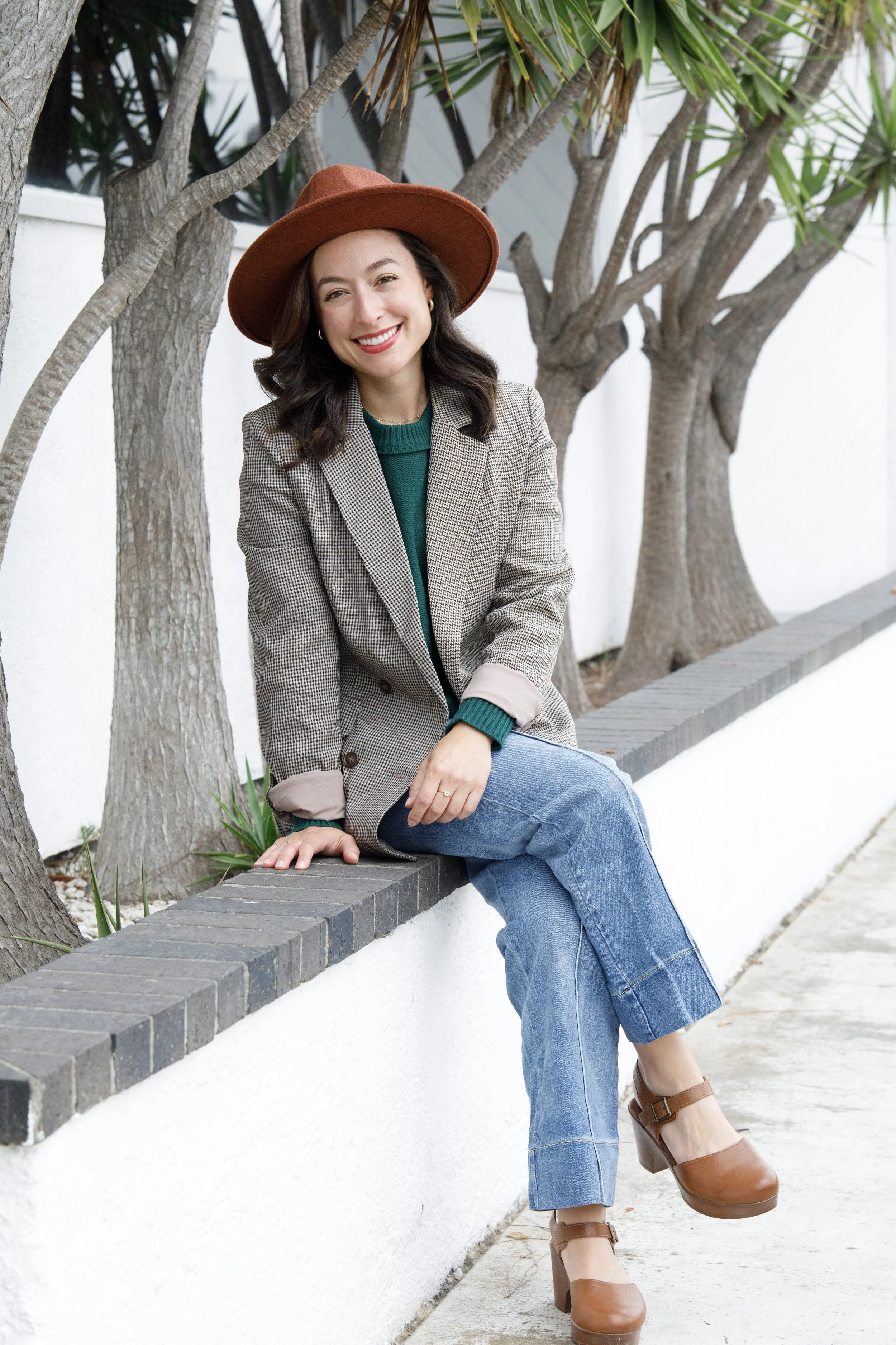 A young woman with dark hair and a bright smile sitting on a white brick ledge in front of trees, wearing a brown hat, plaid blazer, green sweater, blue jeans, and brown shoes.