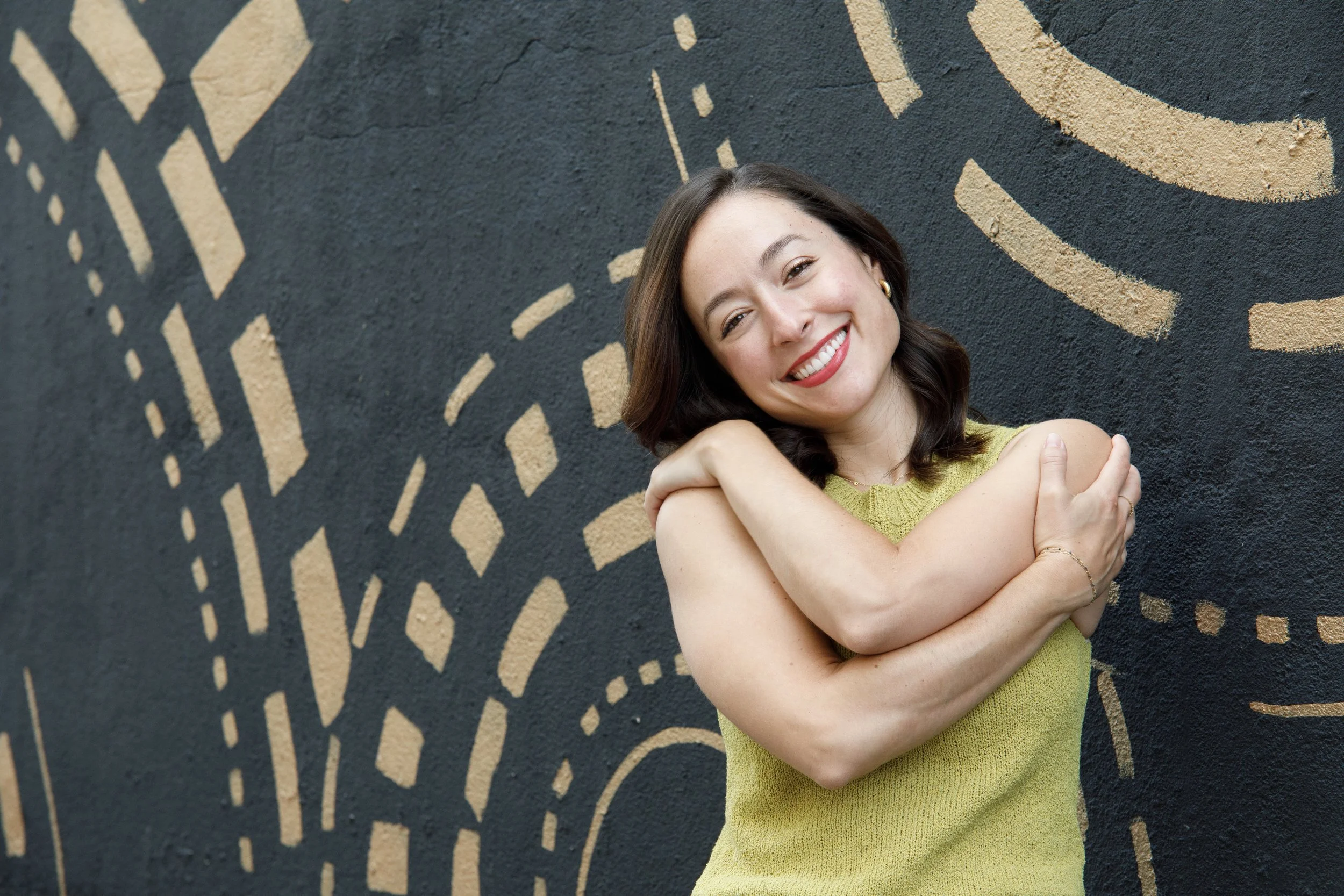 Young woman with shoulder-length dark hair, smiling, wearing a sleeveless yellow top, hugging herself, standing against a dark wall with abstract tan-colored painted lines.