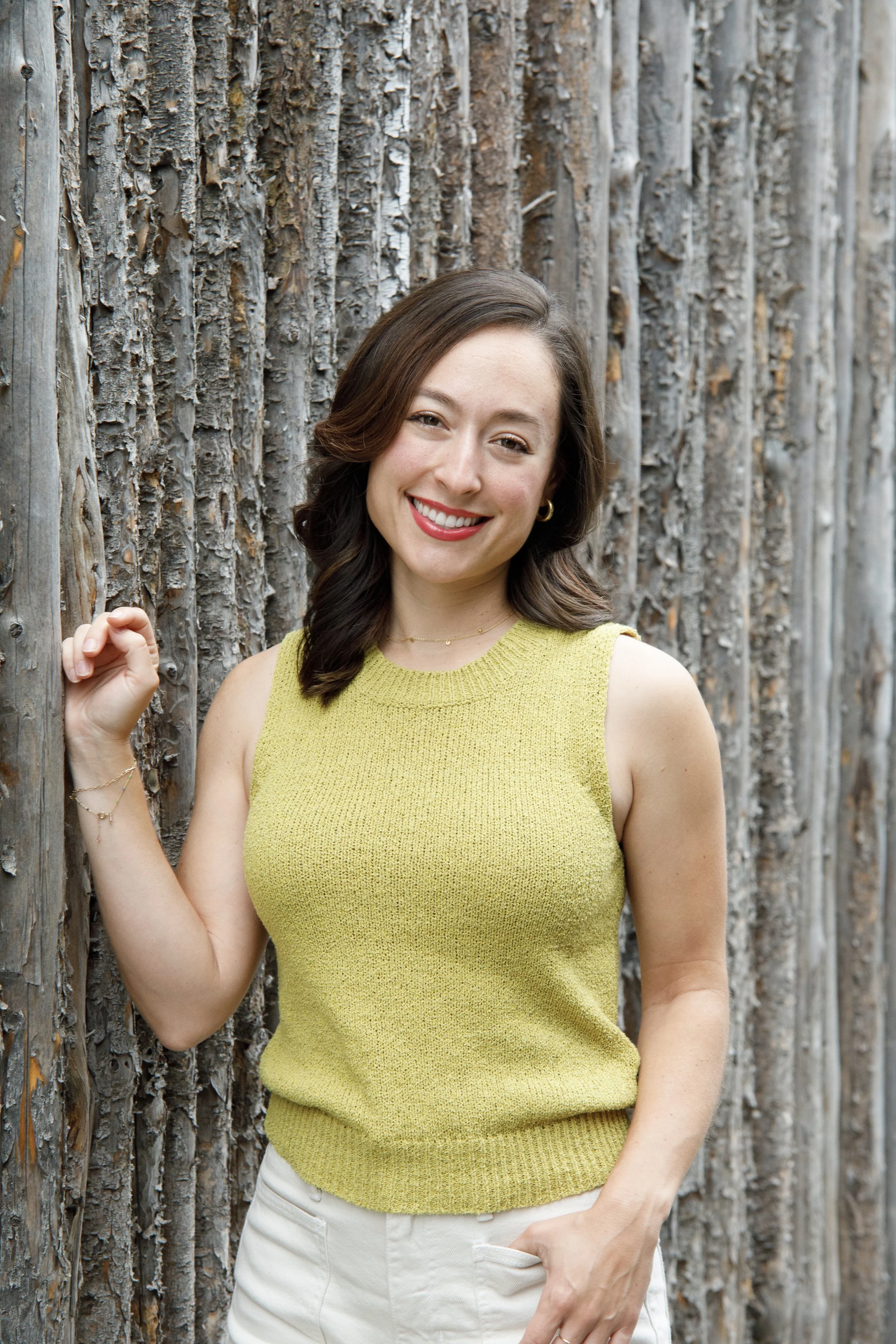 A woman with shoulder-length brown hair, wearing a sleeveless yellow top, smiling and standing against a background of wooden logs.