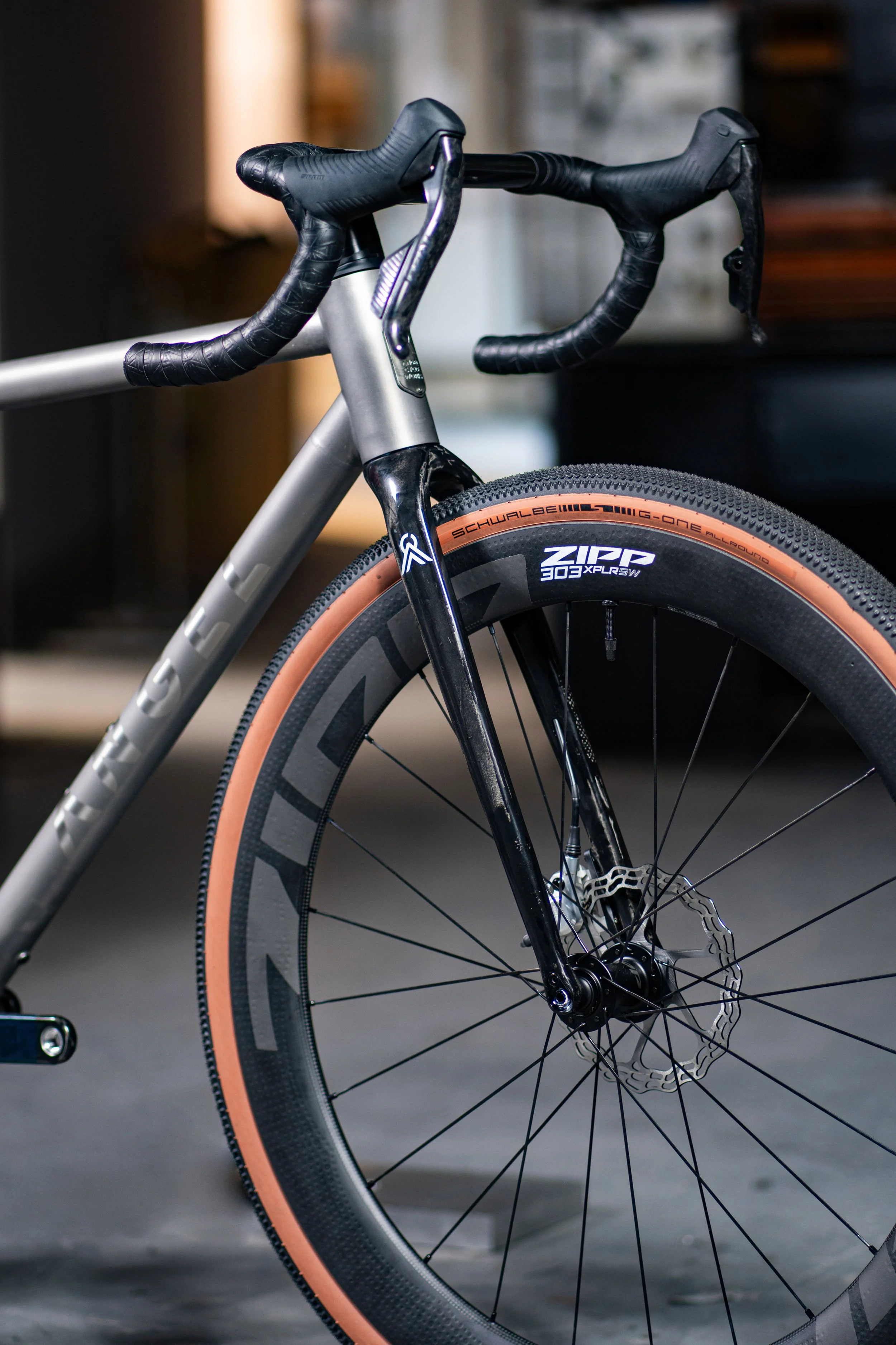 Close-up of a modern road bicycle's front wheel, fork, and handlebars. The black front wheel has a tan sidewall tire, and the bike features disc brakes. The frame is metallic gray, with the brand name partially visible.