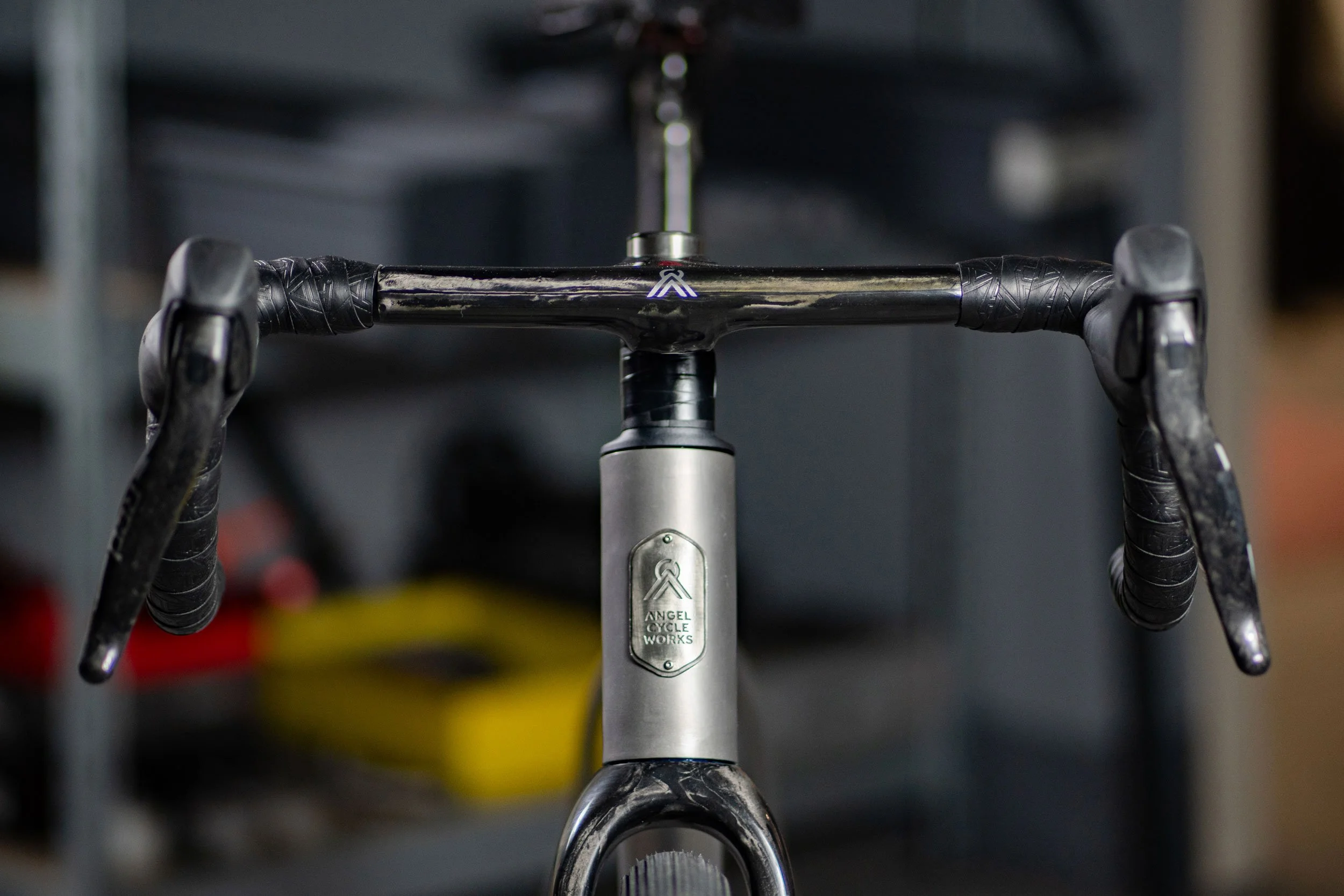 Close-up of a bicycle's handlebar and stem with a silver Angel Cycle Works logo, set in a workshop environment.