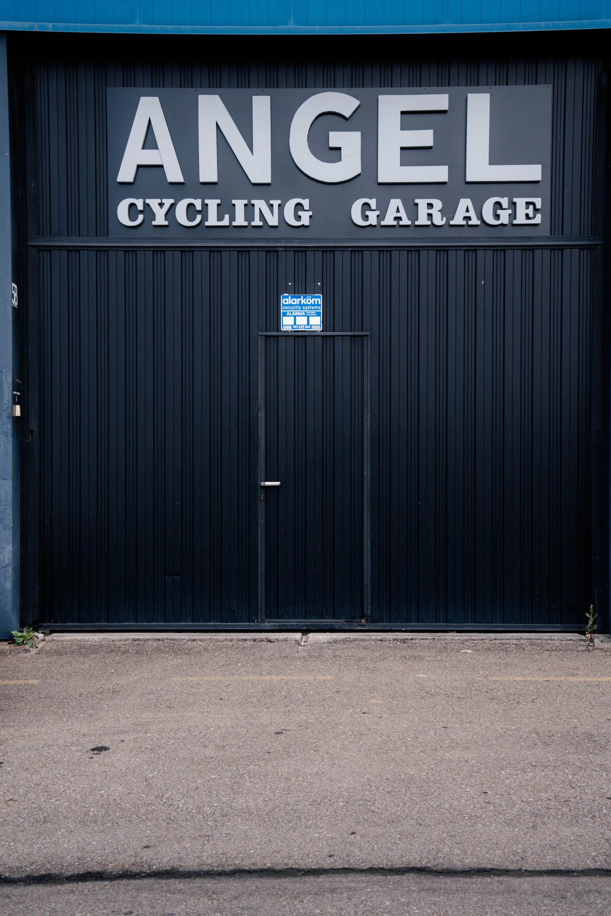 Una cortina de garaje negra con un cartel que dice 'Angel Cycling Garage' en letras grandes y blancas. Debajo, un cartel de alarma alarkom en color azul y blanco.