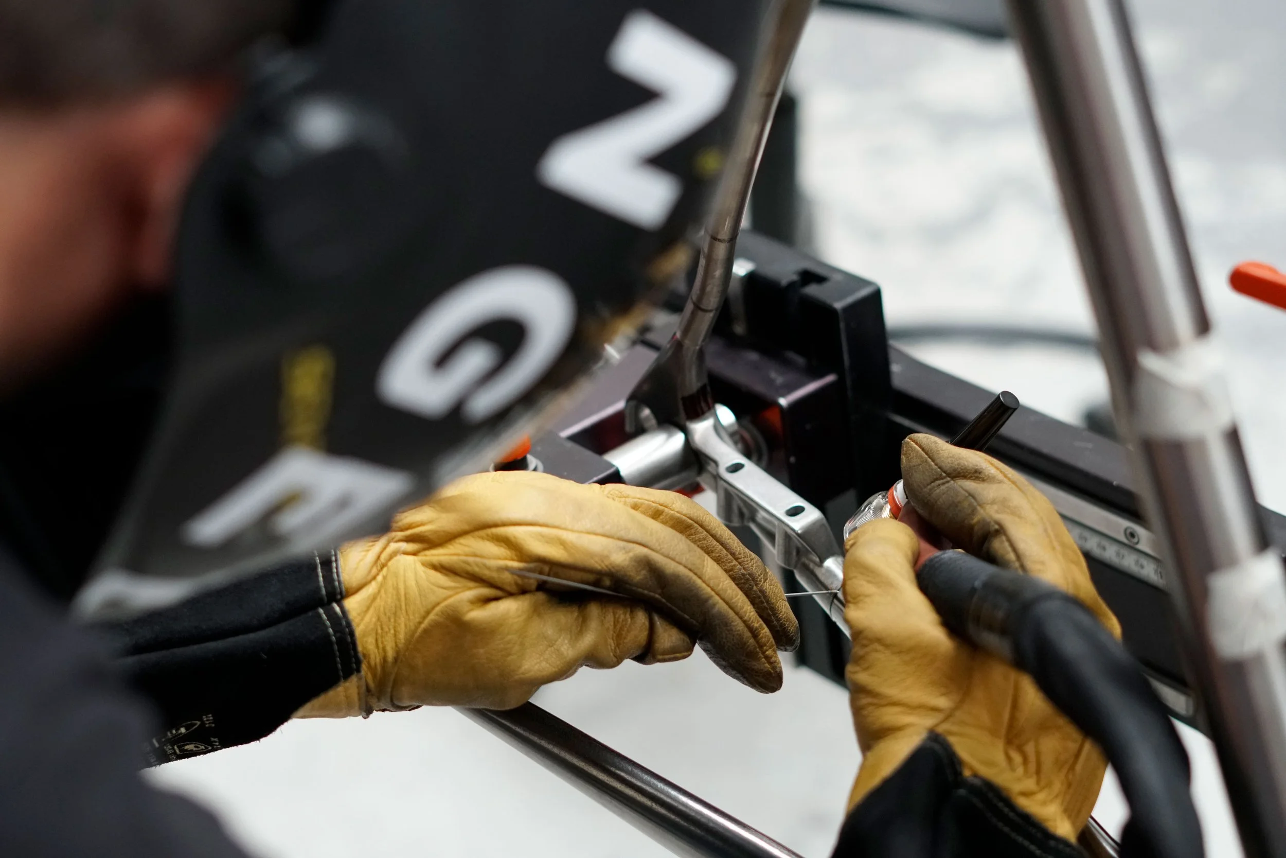 A race car driver is working on a vehicle, wearing yellow gloves and a black helmet, using a tool to adjust a component in the cockpit area.