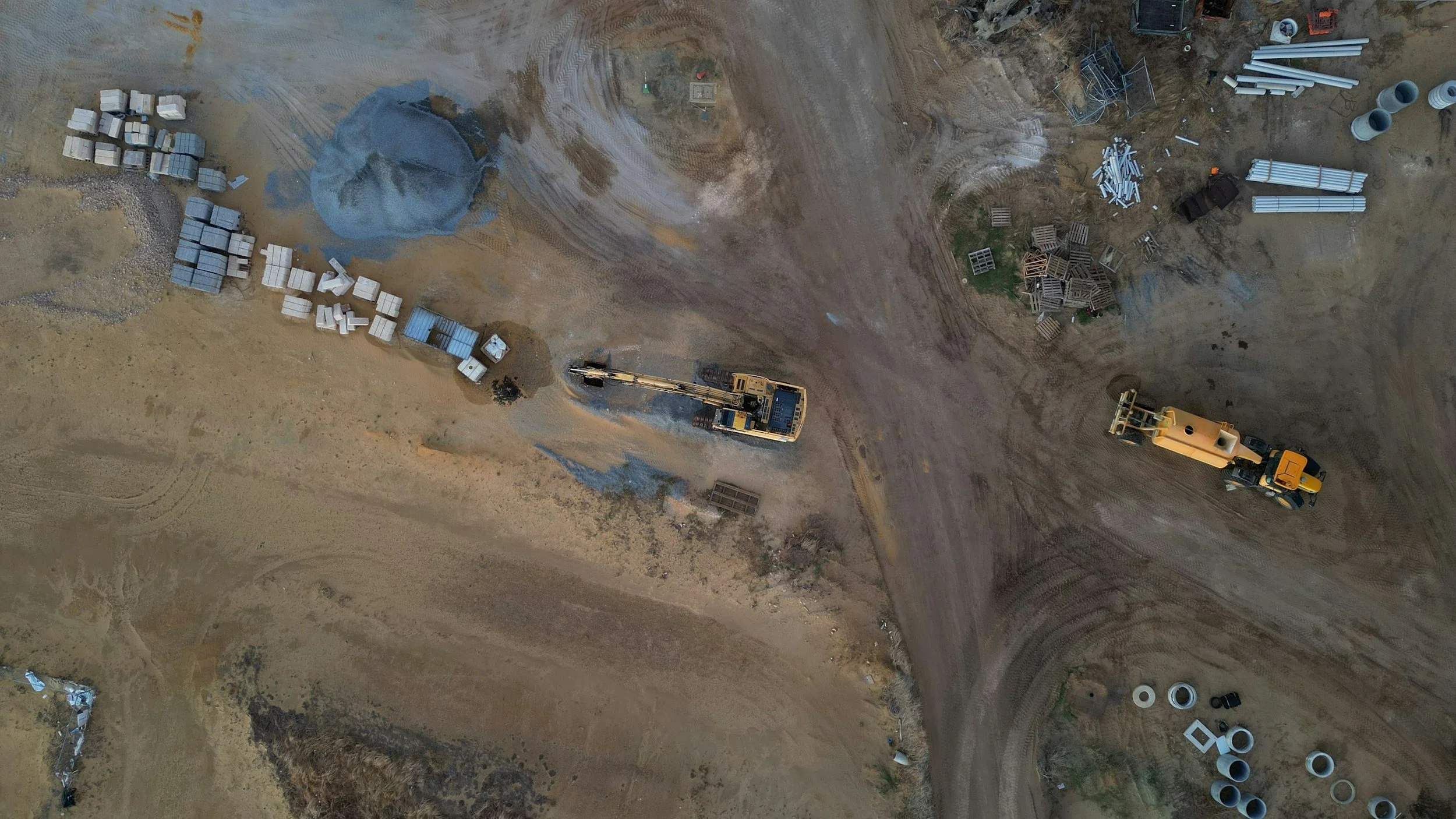 An aerial view of a construction site with machinery, piles of pipes, and building materials on bare earth.
