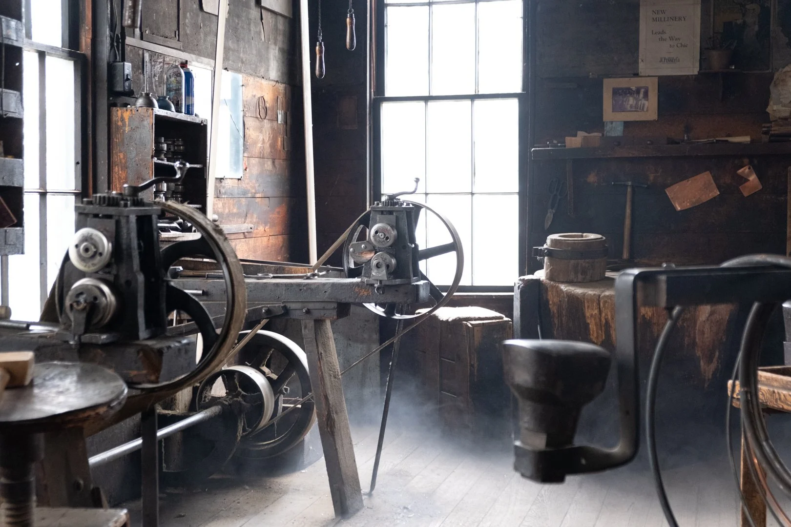 An old workshop with vintage equipment, including a large spinning machine, wooden workbenches, and tools, illuminated by natural light from a window.