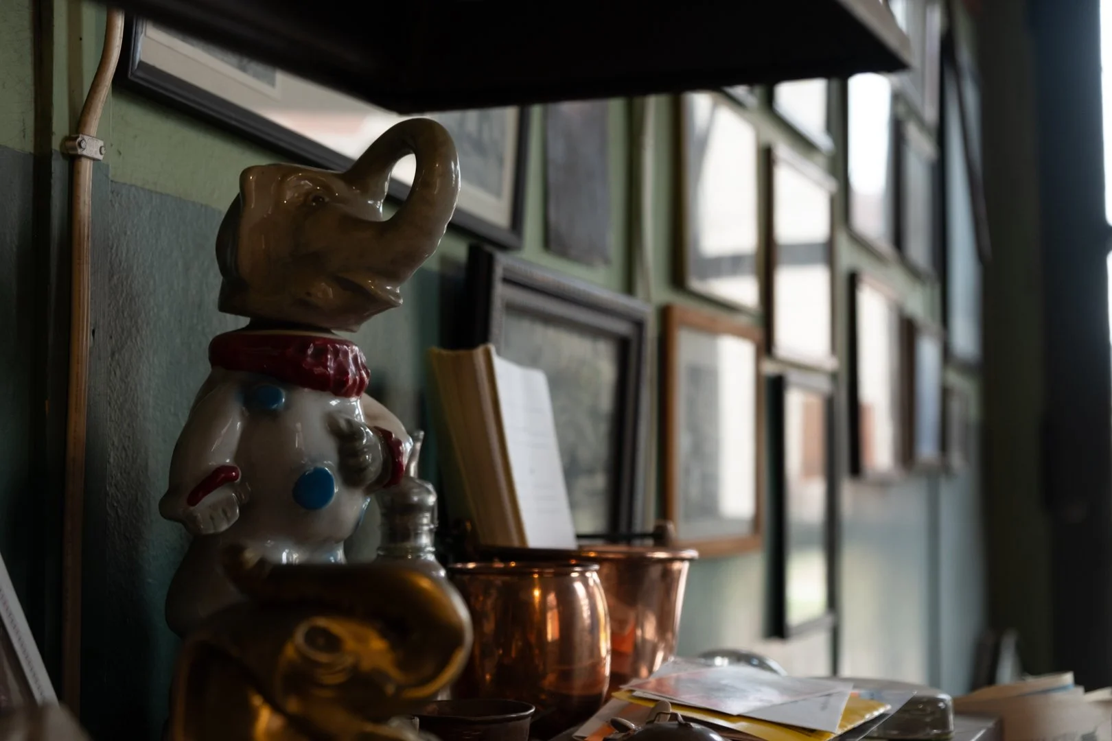 Ceramic elephant figurine wearing a chef's coat with utensils, on a shelf in a room with framed pictures and documents.
