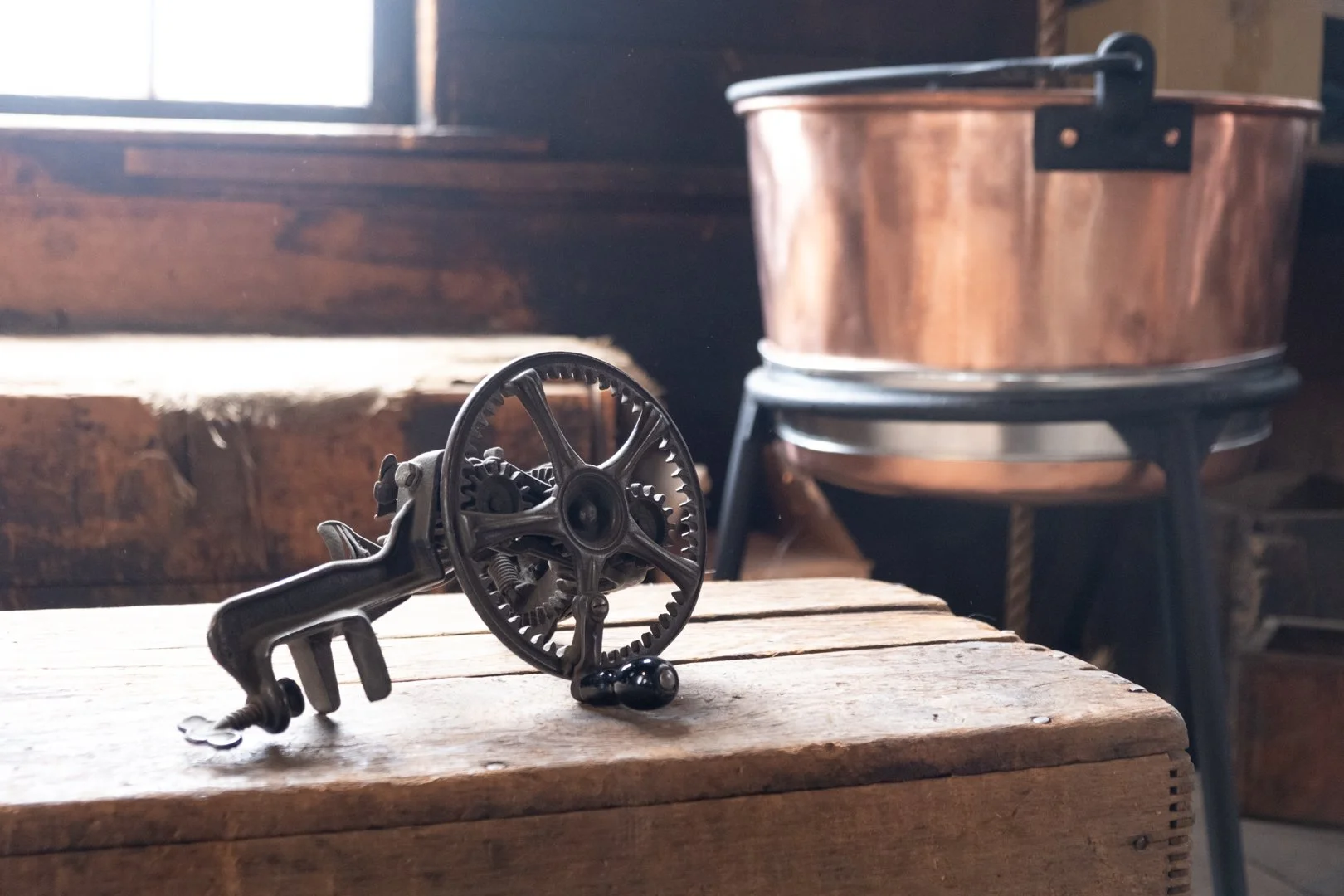 Old fishing reel on a wooden table with a copper cooking pot in the background near a window.