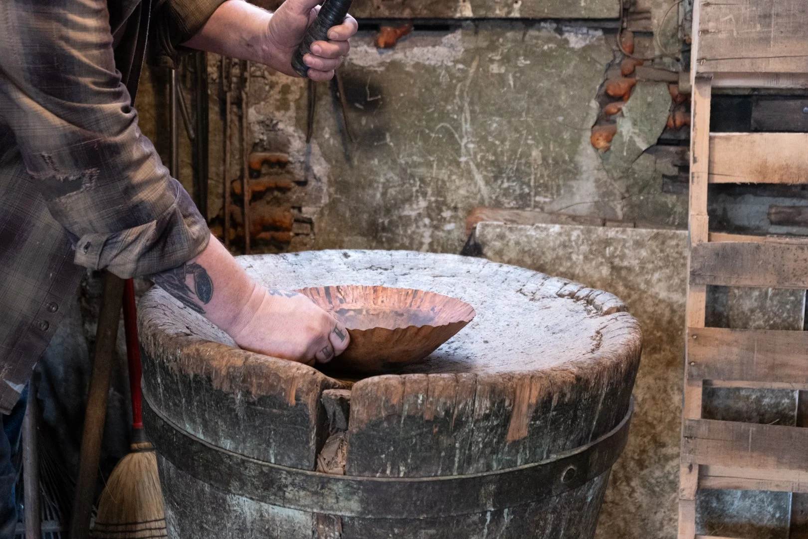 A person using a hammer to shape a metal bowl in a rustic workshop.