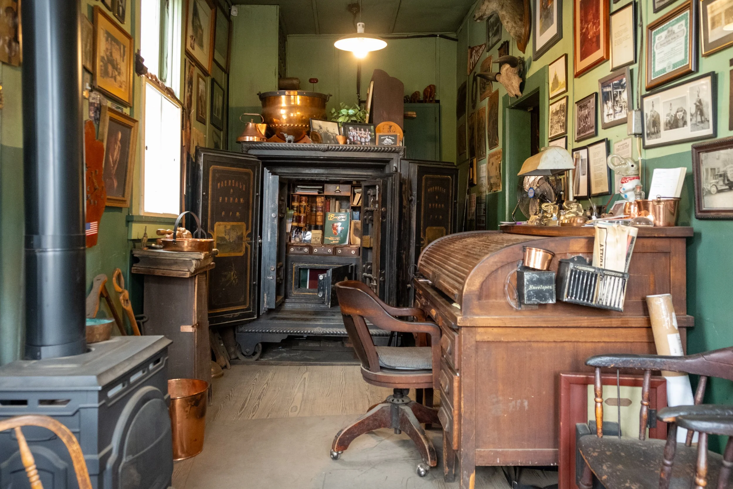 An eclectic vintage room filled with antique furniture, framed photos on green walls, and various old items including a wooden desk, rolling chair, metal safe, and a stove.