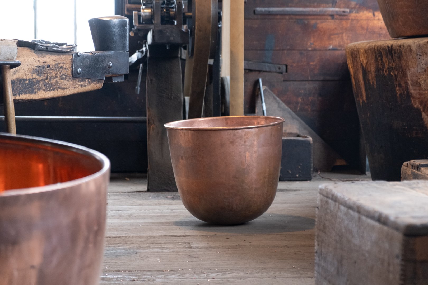 A copper bowl on a wooden floor in a rustic workshop with wooden and metal tools and furniture.