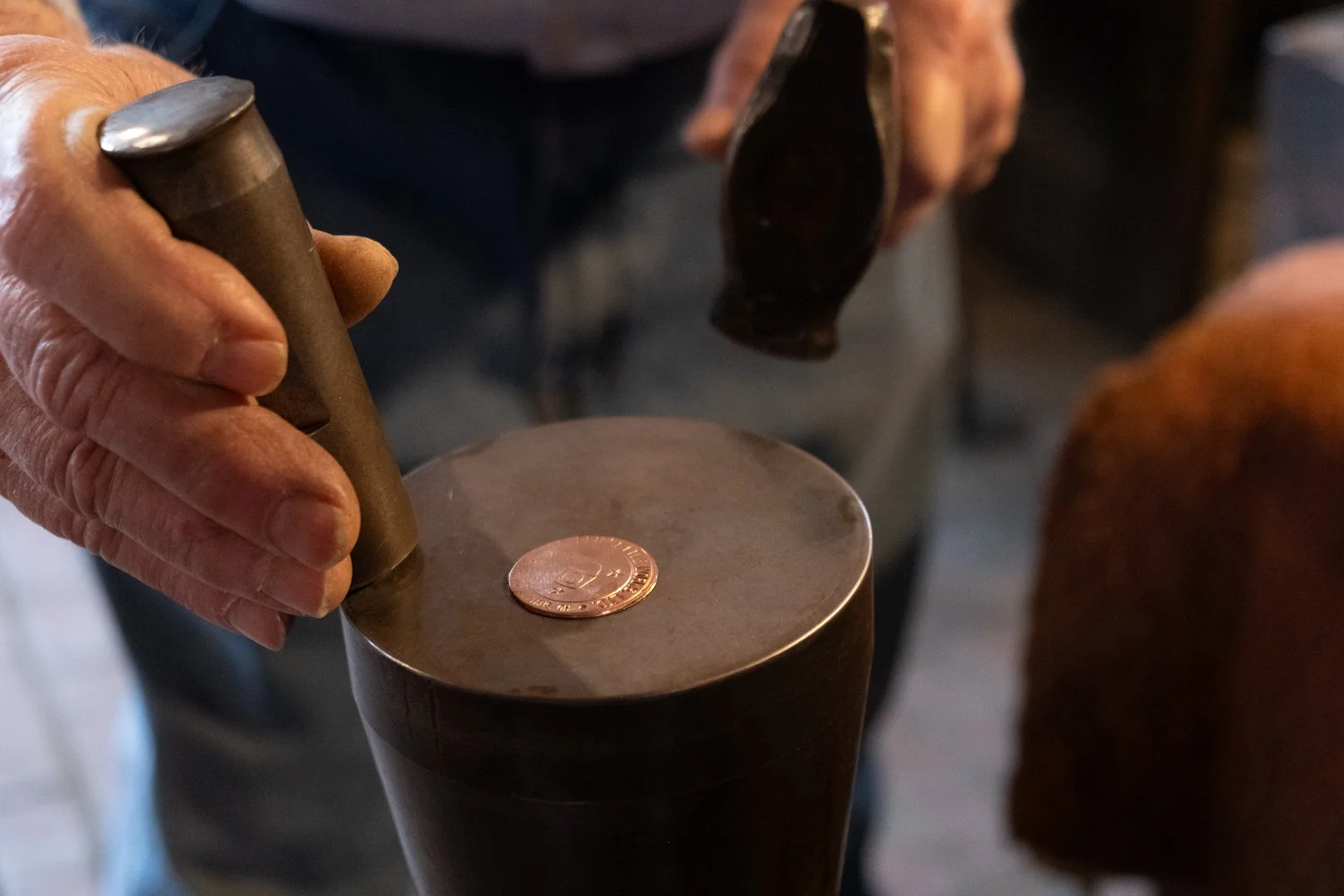 A person using a hammer to strike a metal stamp on a coin to imprint a design, with a copper penny in the background.