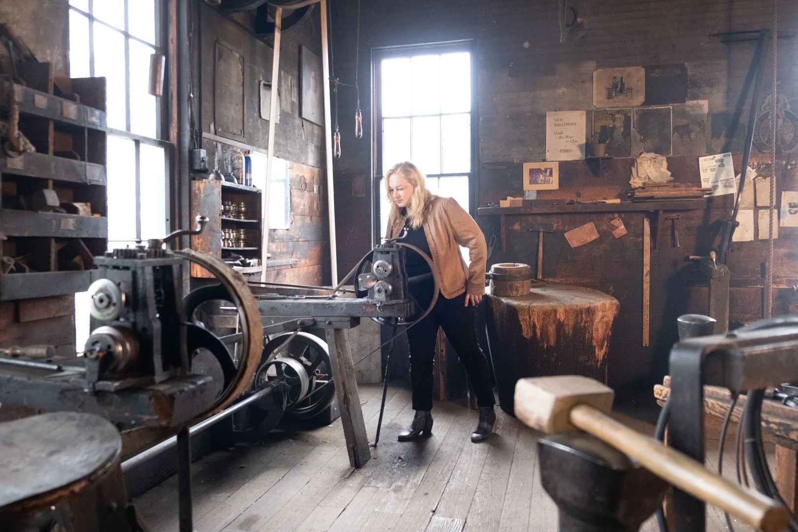 A woman is working with machinery inside an old workshop or blacksmith's shop with wooden walls and large windows.