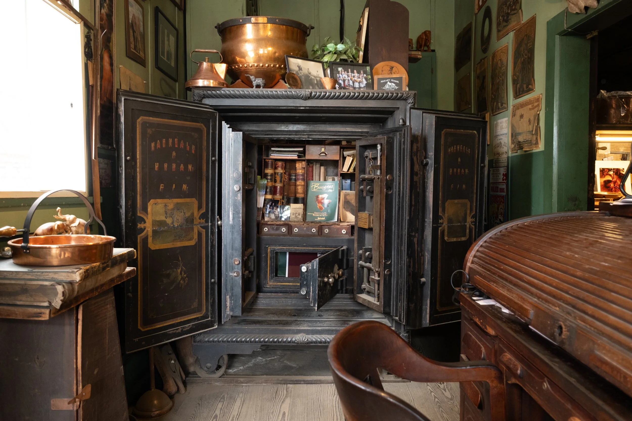 An antique safe vault with its door open displaying books and boxes inside. Surrounding objects include copper cookware, wooden furniture, and framed pictures on green walls.