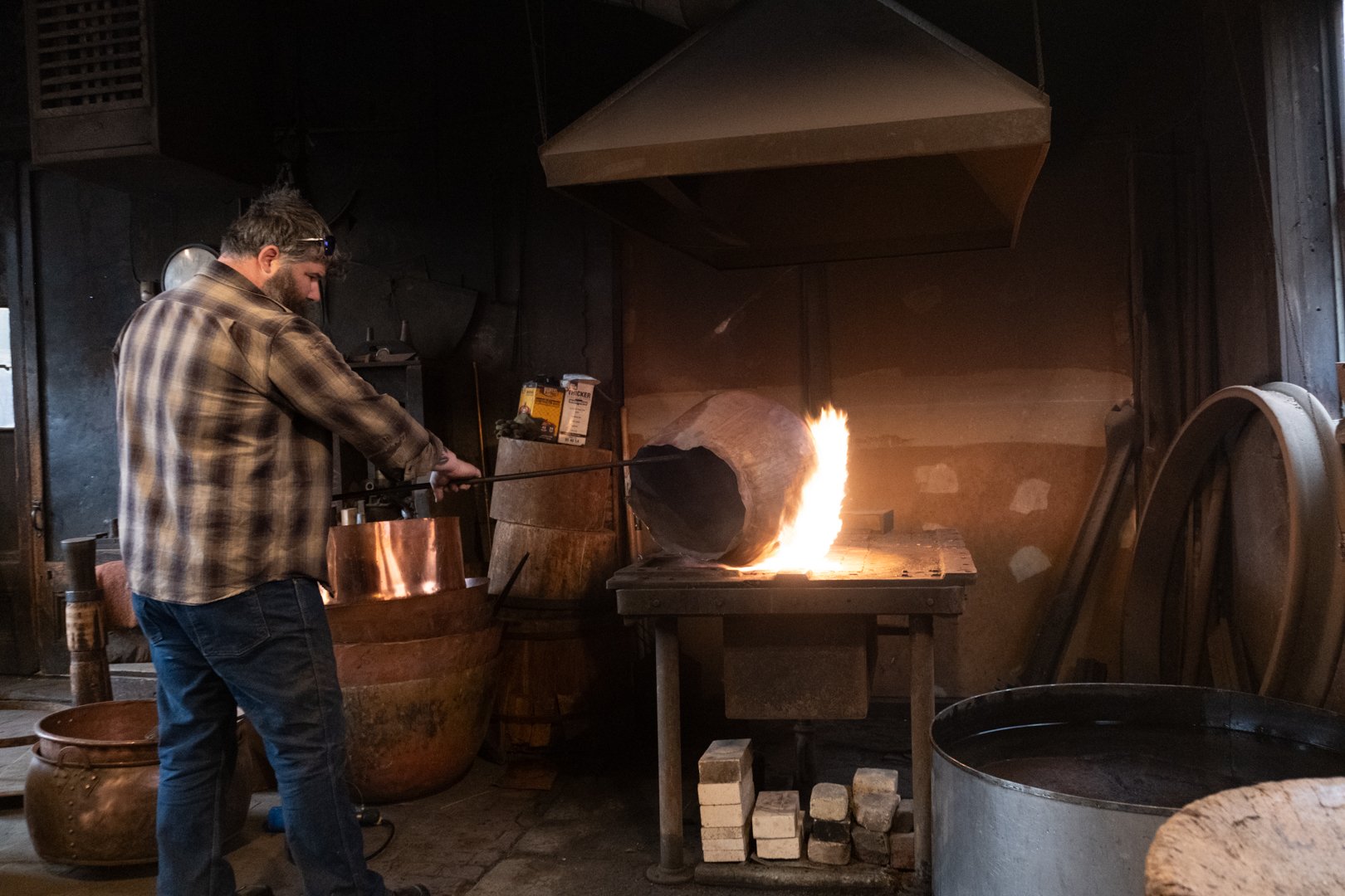 Blacksmith working with fire in a forge, wearing a plaid shirt and safety glasses, inside a workshop with various tools and equipment.