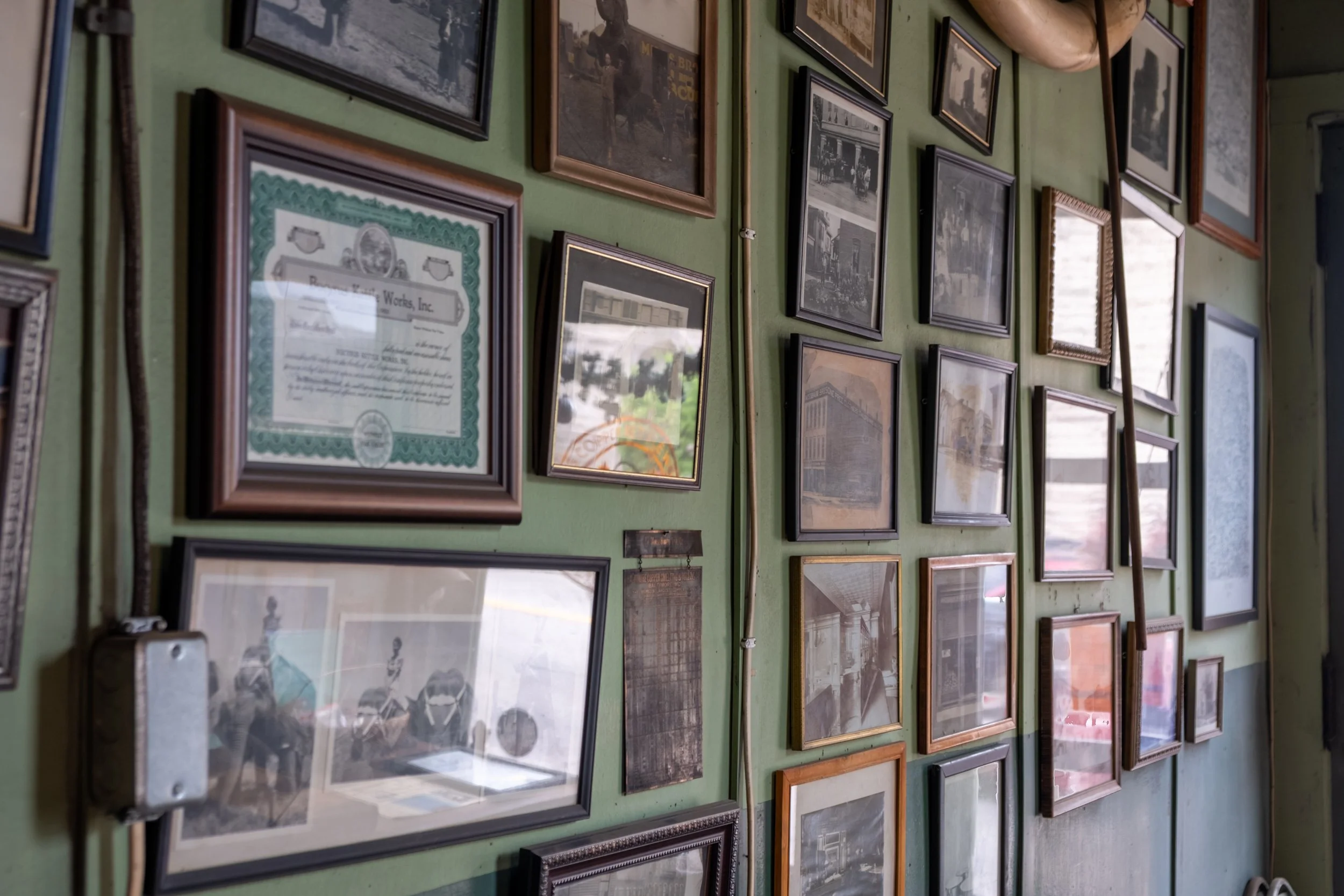 A green wall decorated with numerous framed photographs and documents. The images mostly appear to be vintage black-and-white photos, and the documents include a visible certificate with a decorative border.
