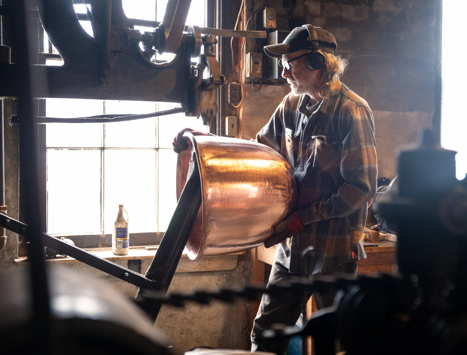A man in a plaid shirt, cap, glasses, and headphones physicalizes a large copper object in a workshop with a window in the background.
