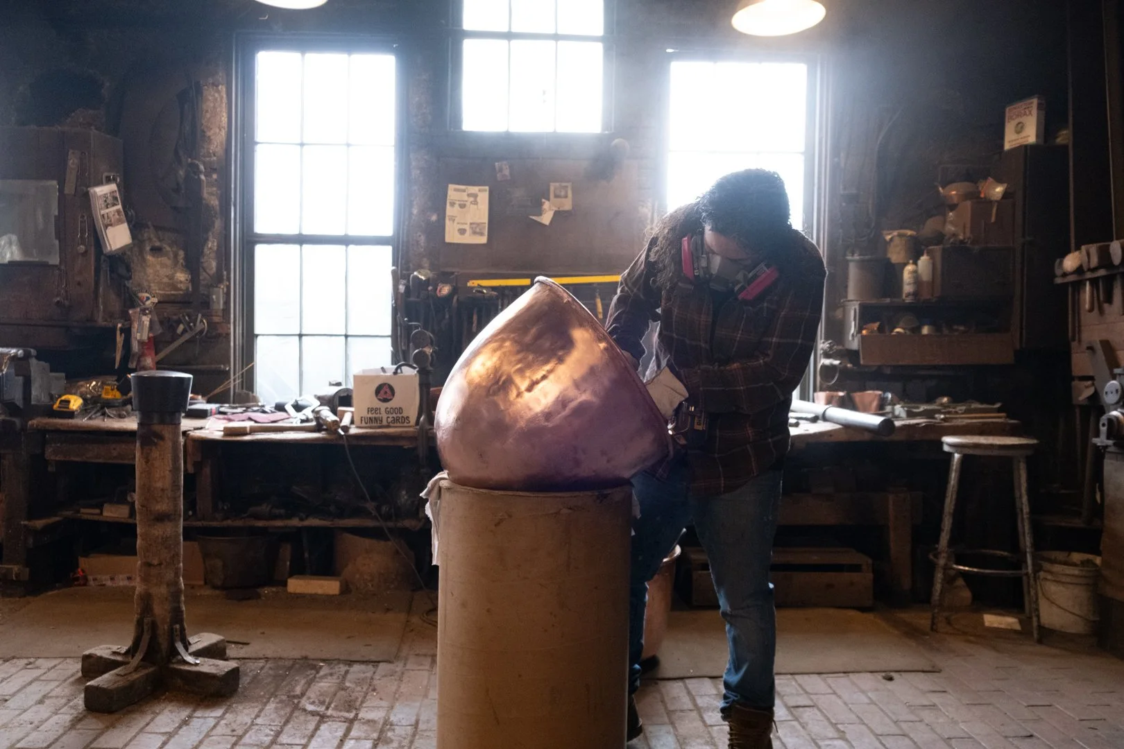 A person working on a large metallic object in a workshop, wearing safety goggles and a respirator mask, with tools and supplies on shelves and a workbench behind them.