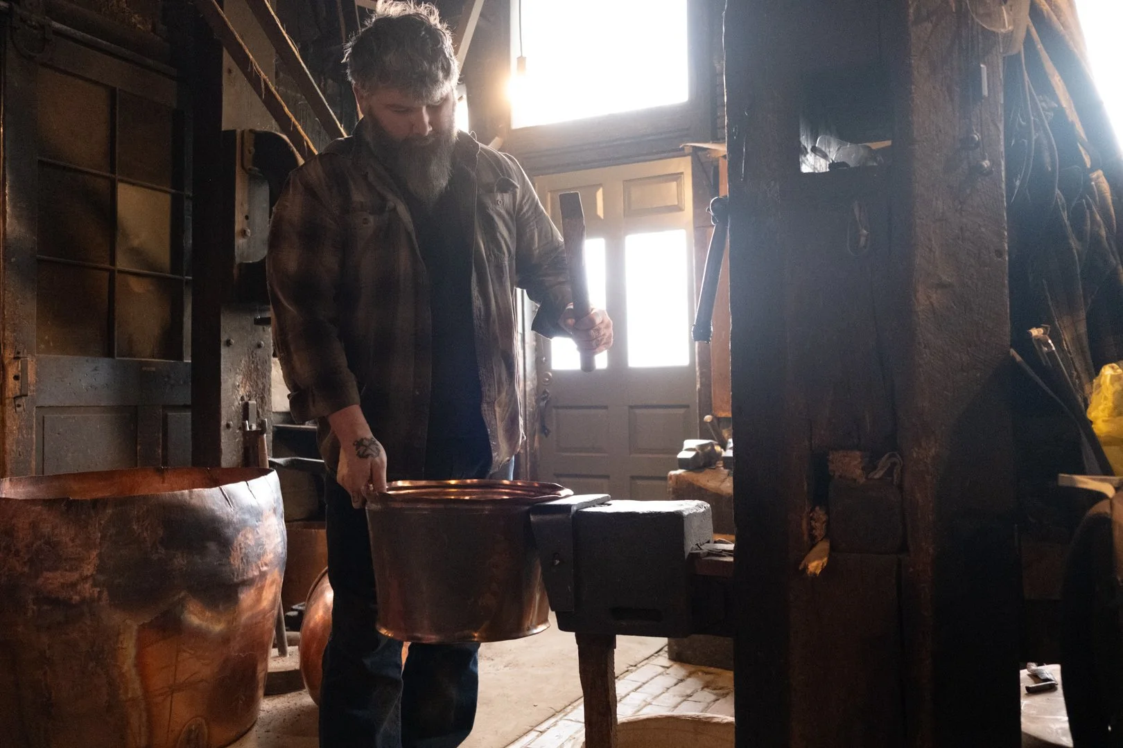 A man with a beard standing in a rustic workshop, holding a hammer and working on a copper bowl or similar object.