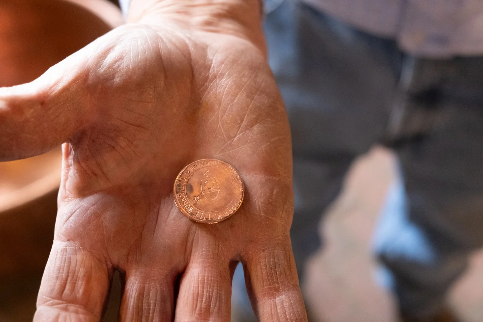 A close-up of a person's hand showing the back, with a penny resting on the palm. The hand appears aged with visible wrinkles and veins, and the person is wearing a gray shirt and blue jeans.
