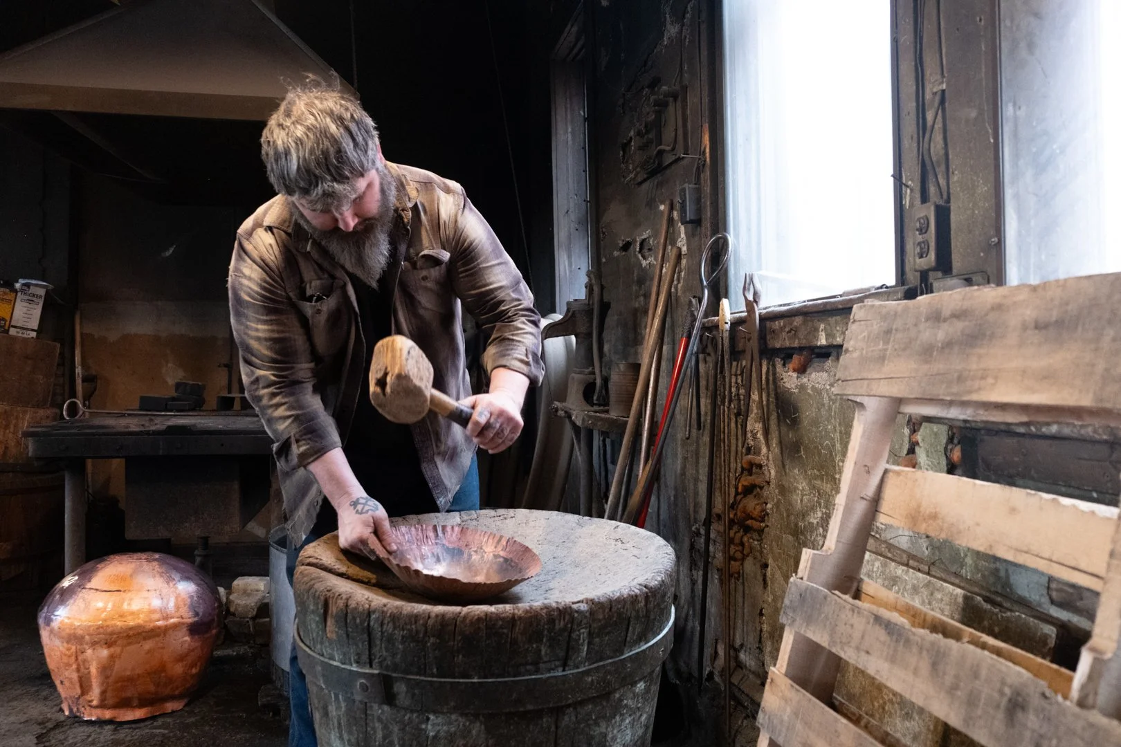 A man with a beard, wearing a brown shirt, is using a hammer on a carved wooden bowl in a workshop with tools and a window.