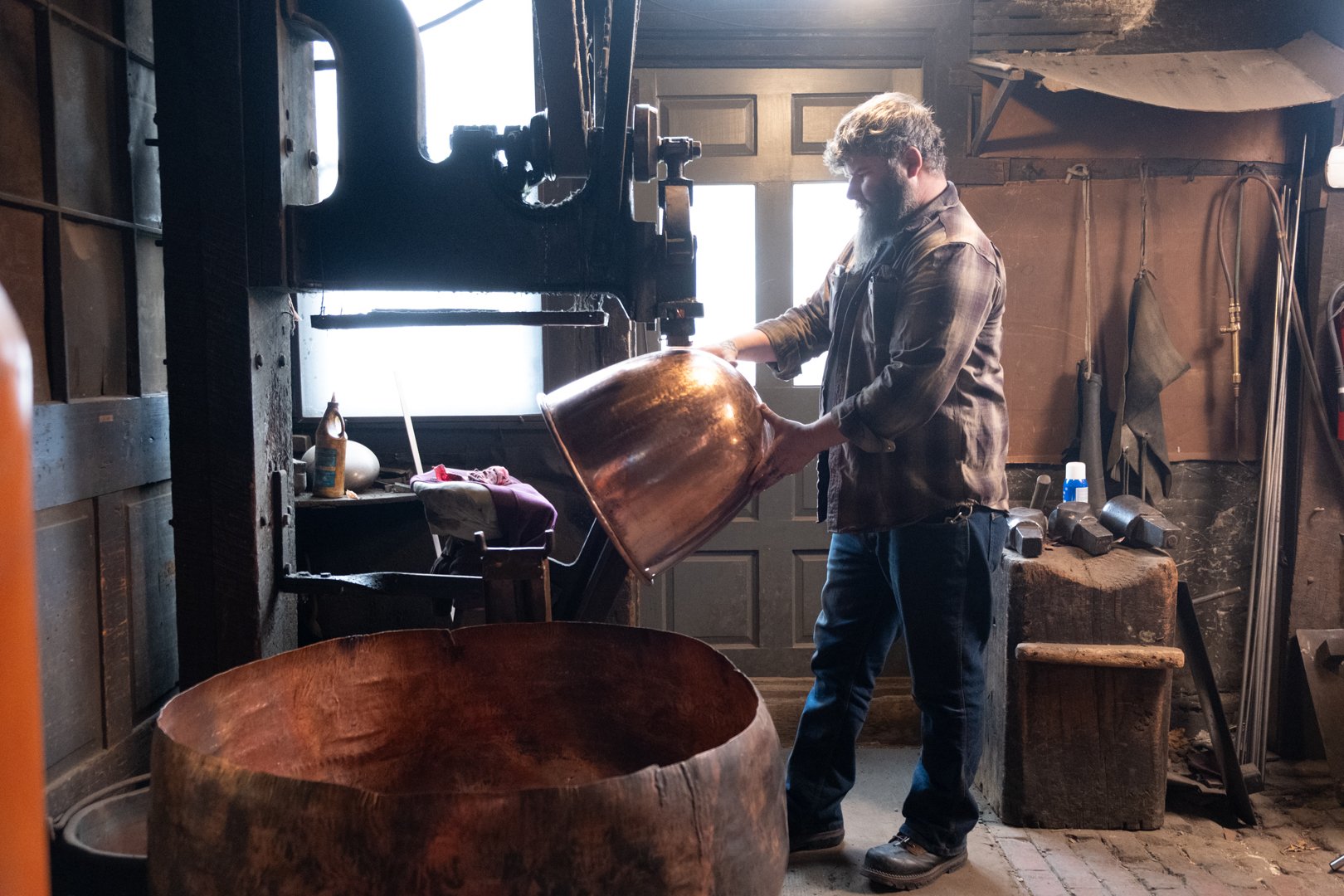 A man working in a woodworking shop, pouring a large copper or bronze bowl into a wooden vessel.