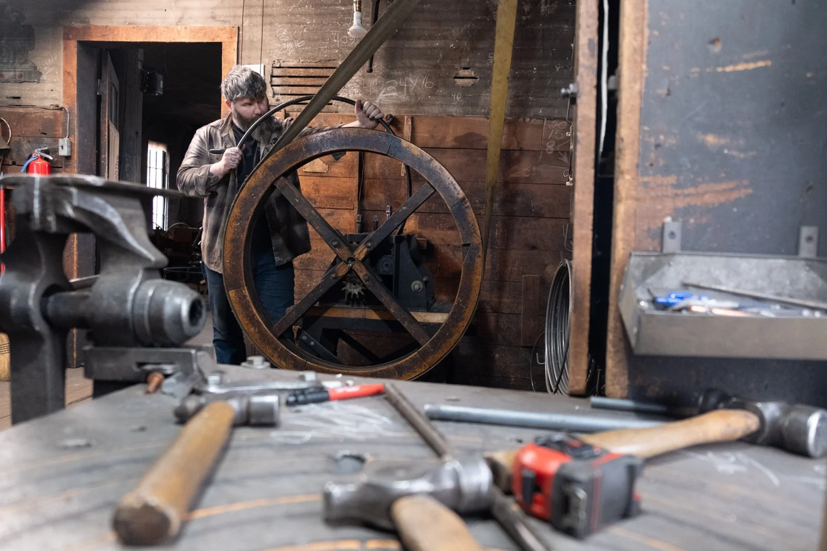 A man working in a workshop with tools scattered on a workbench in the foreground, and a large industrial wheel or gear being worked on in the background.