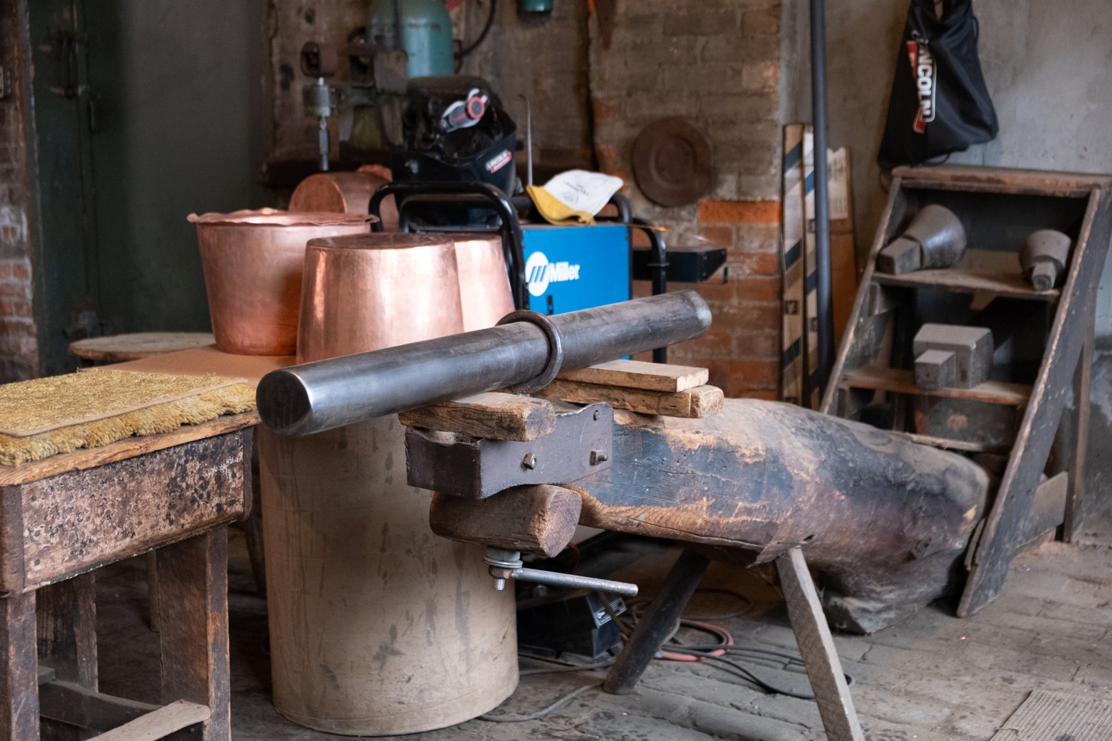 A workshop with metalworking tools, copper vessels, a workbench, a vice, and a wooden shelf storing metal pieces, set against a brick wall.