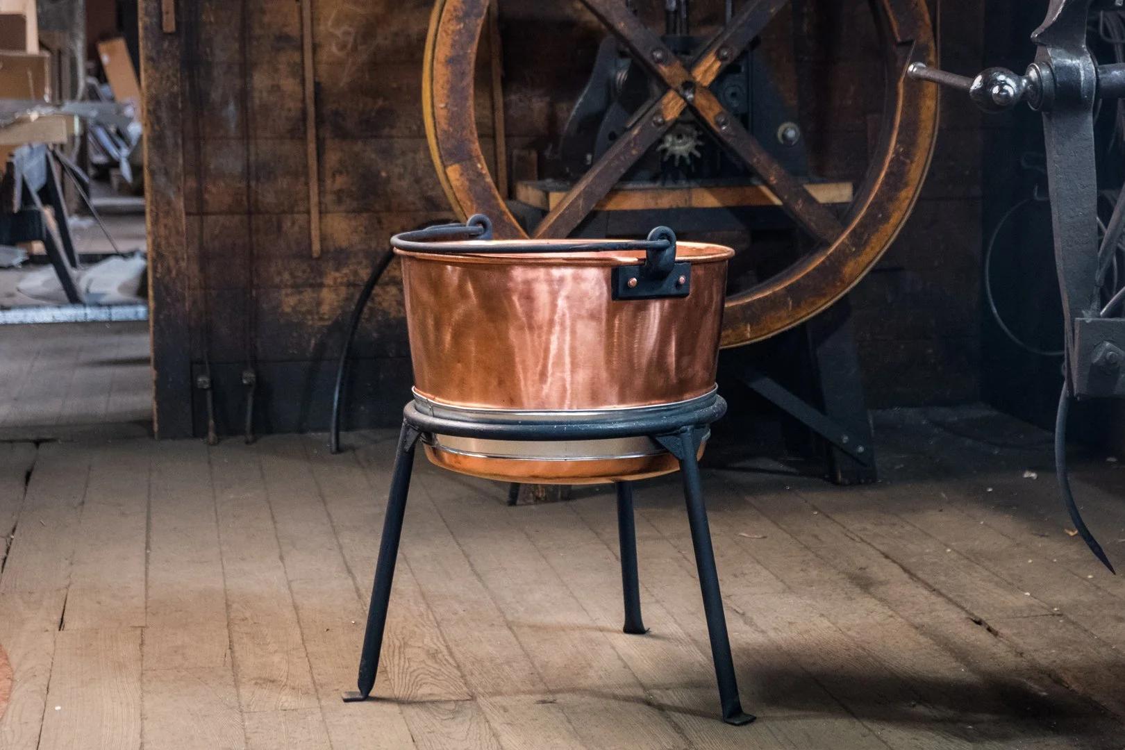 Copper brewing kettle on a black metal stand inside a rustic workshop.