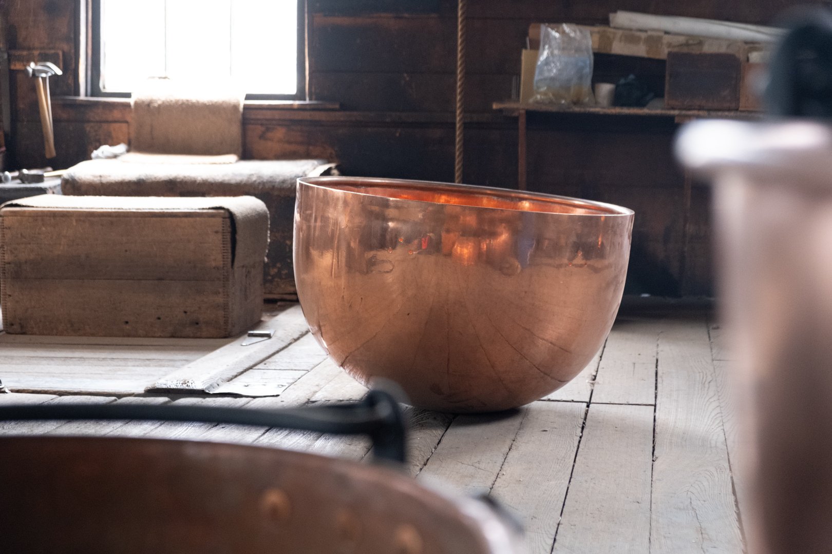 A large copper singing bowl on a wooden floor in a rustic room with wooden walls, a window, and vintage furniture.