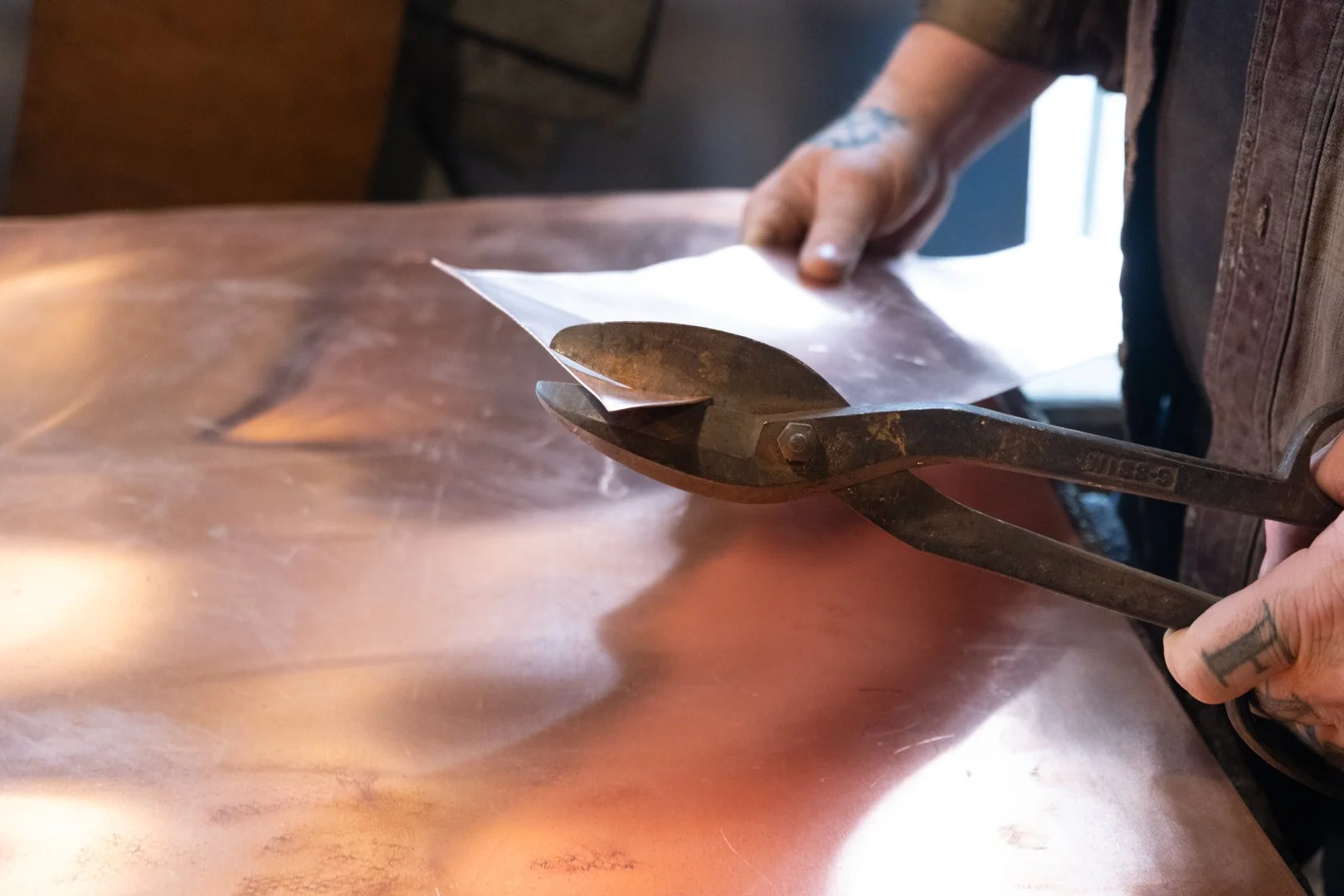 Hands holding large metal shears cutting a piece of thin, shiny copper sheet.