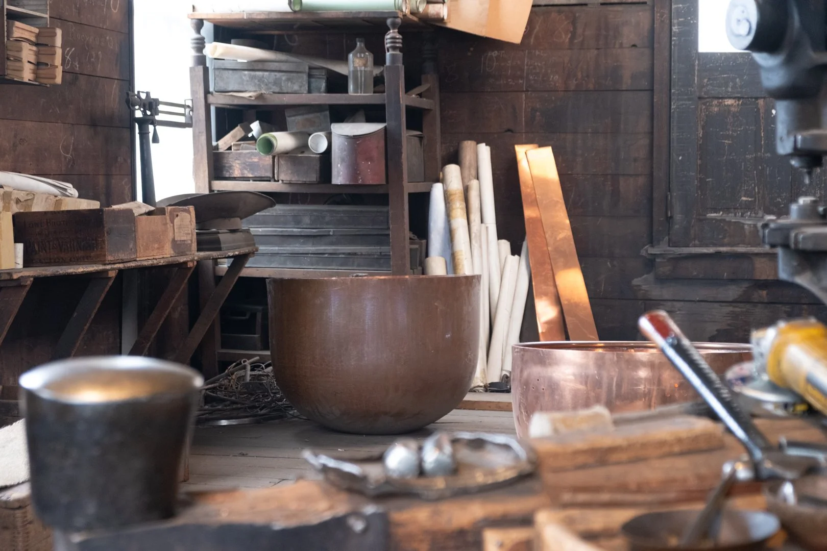 A woodworking workshop with various tools, wooden bowls, metal sheets, and supplies on shelves and workbenches.
