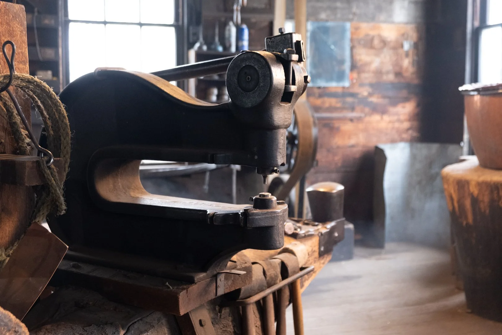 An antique black cast iron sewing machine on a wooden workbench in a rustic workshop with wooden walls and large windows.
