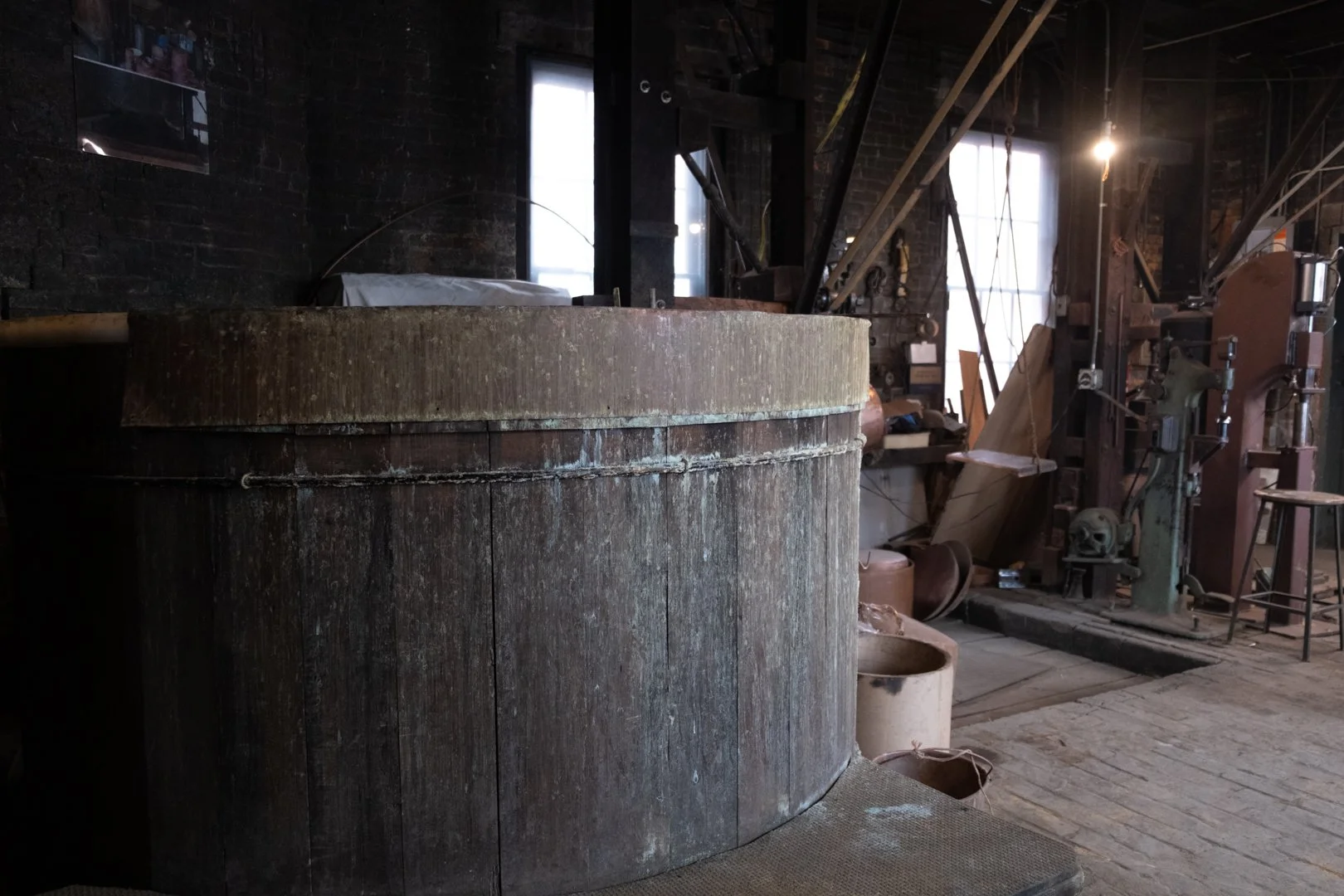Interior of a blacksmith shop showcasing an anvil with a rounded, aged metal surface, various tools, and equipment related to metalworking, with wooden floors and brick walls illuminated by natural light.