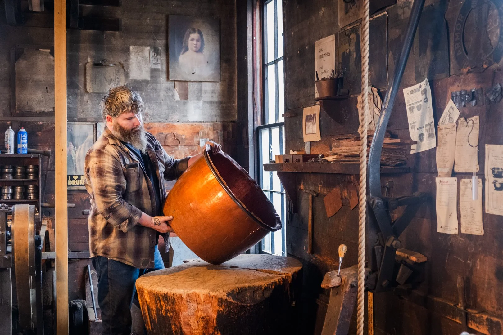 A bearded man in a plaid shirt pouring a large wooden bucket onto a chopping block in a rustic workshop with wood-paneled walls, framed photos, and various tools and papers.