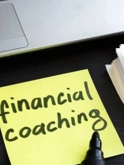 Une feuille jaune avec l'inscription 'financial coaching' écrite à la main, sur un bureau avec un ordinateur portable et un livre à proximité.