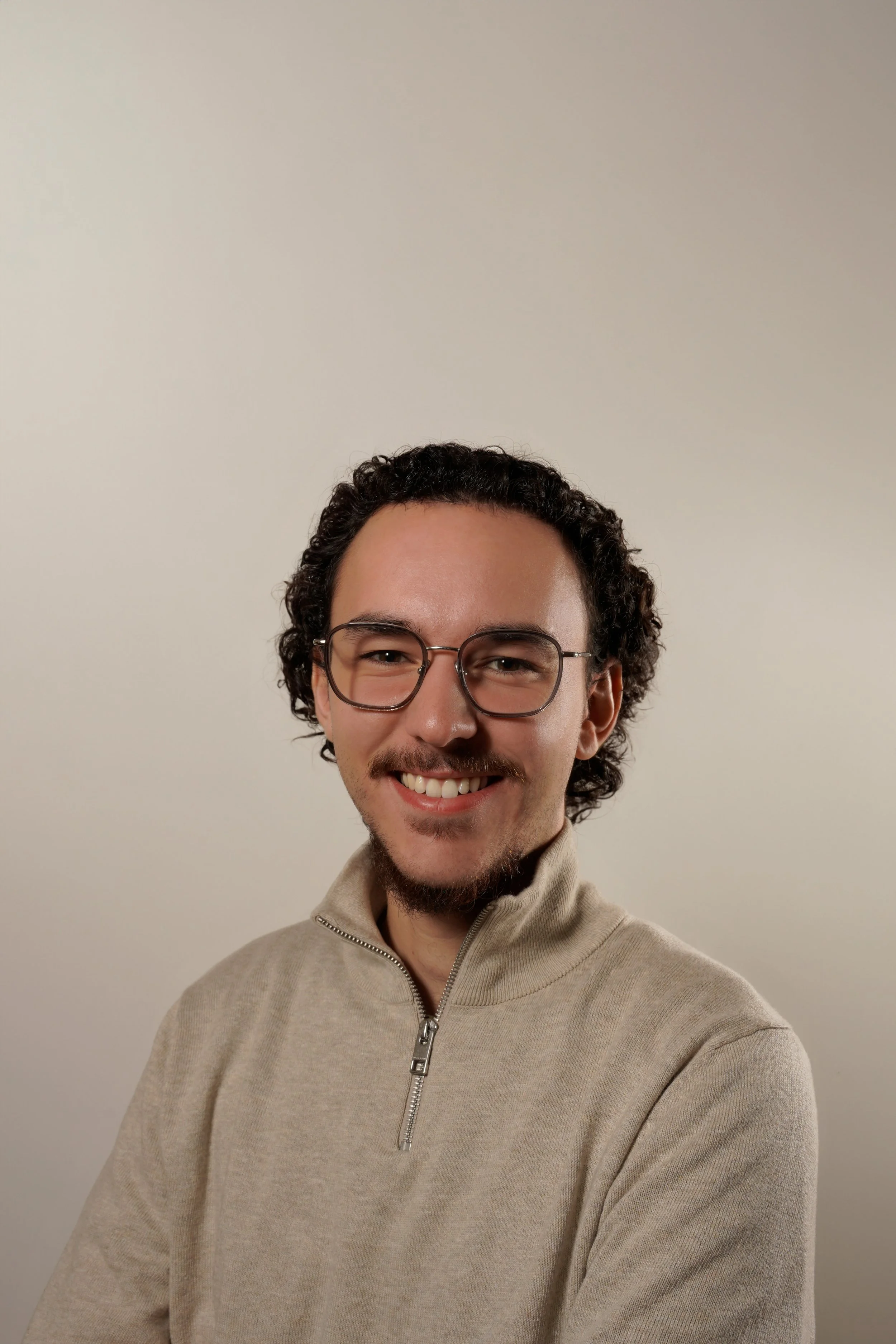 Portrait d'un homme souriant avec lunettes, barbe légère, cheveux bouclés, portant une veste beige à fermeture éclair, sur fond neutre.
