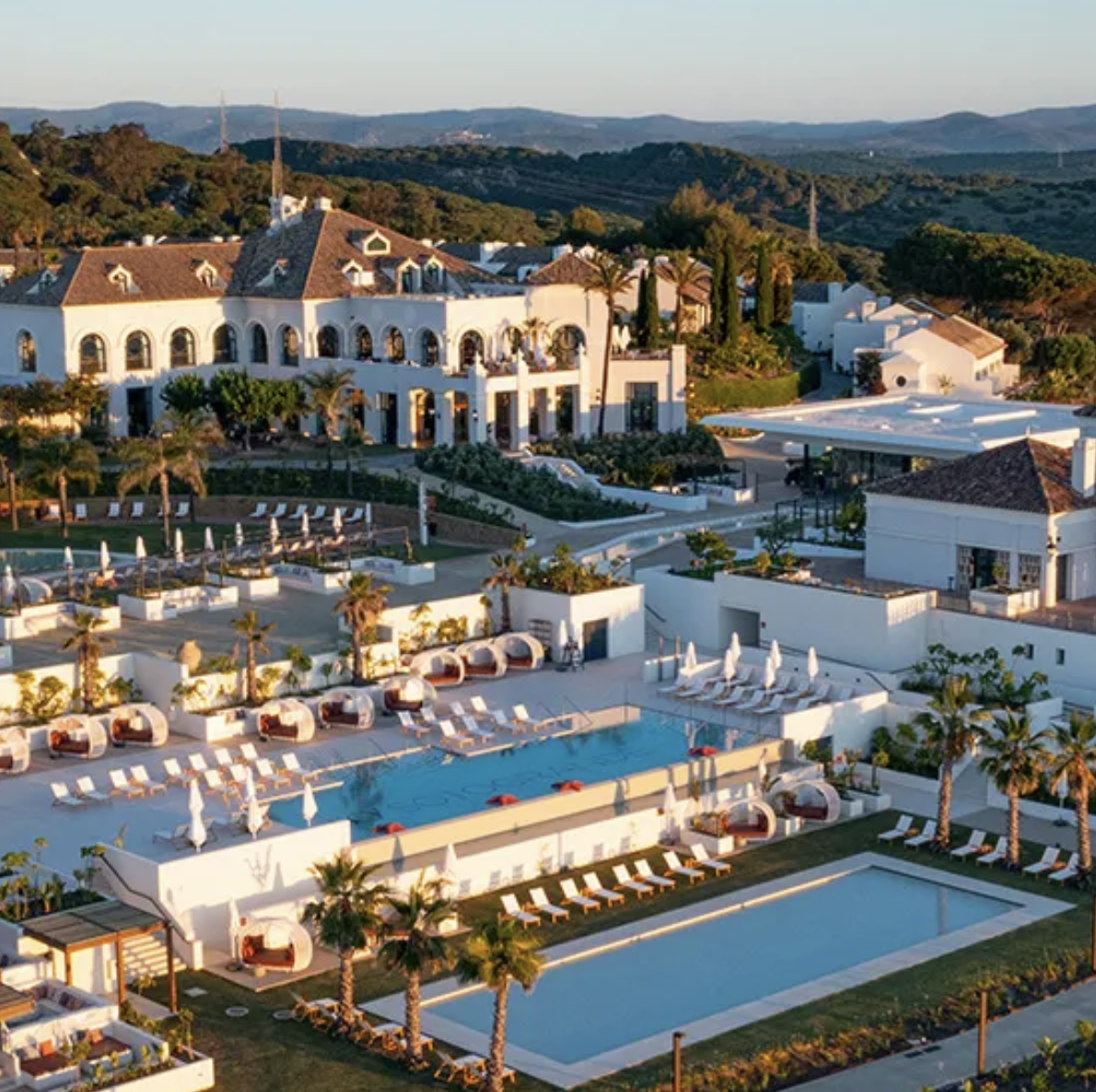 A luxury resort with white buildings, swimming pools, palm trees, lounge chairs, and green hills in the background, taken during sunset. The French Riviera Golf. Caddie & Co. | Discover exclusive golf trips.