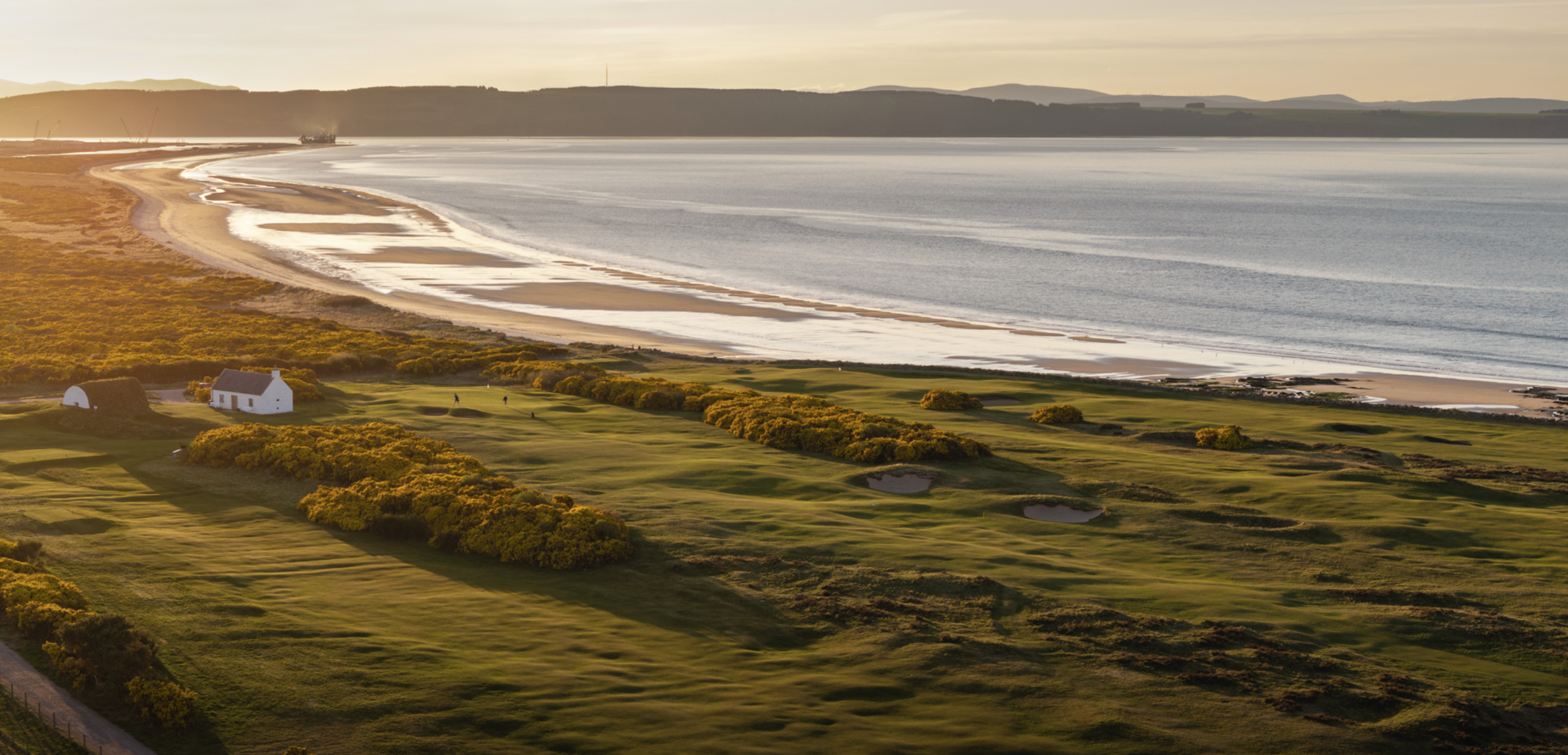 A scenic view of a golf course near a beach at sunset with small white buildings and green fairways. Ireland Golf. Narin. Caddie & Co. | Discover exclusive golf trips.
