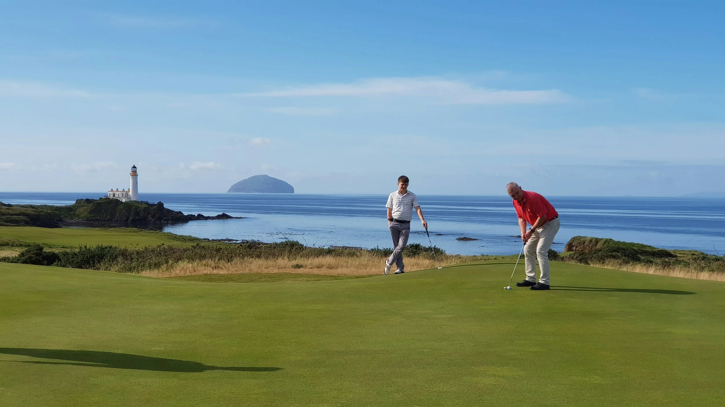 Two men playing golf on a course overlooking a lighthouse and ocean, with an island in the distance. Scotland Golf. Caddie & Co. | Discover exclusive golf trips.