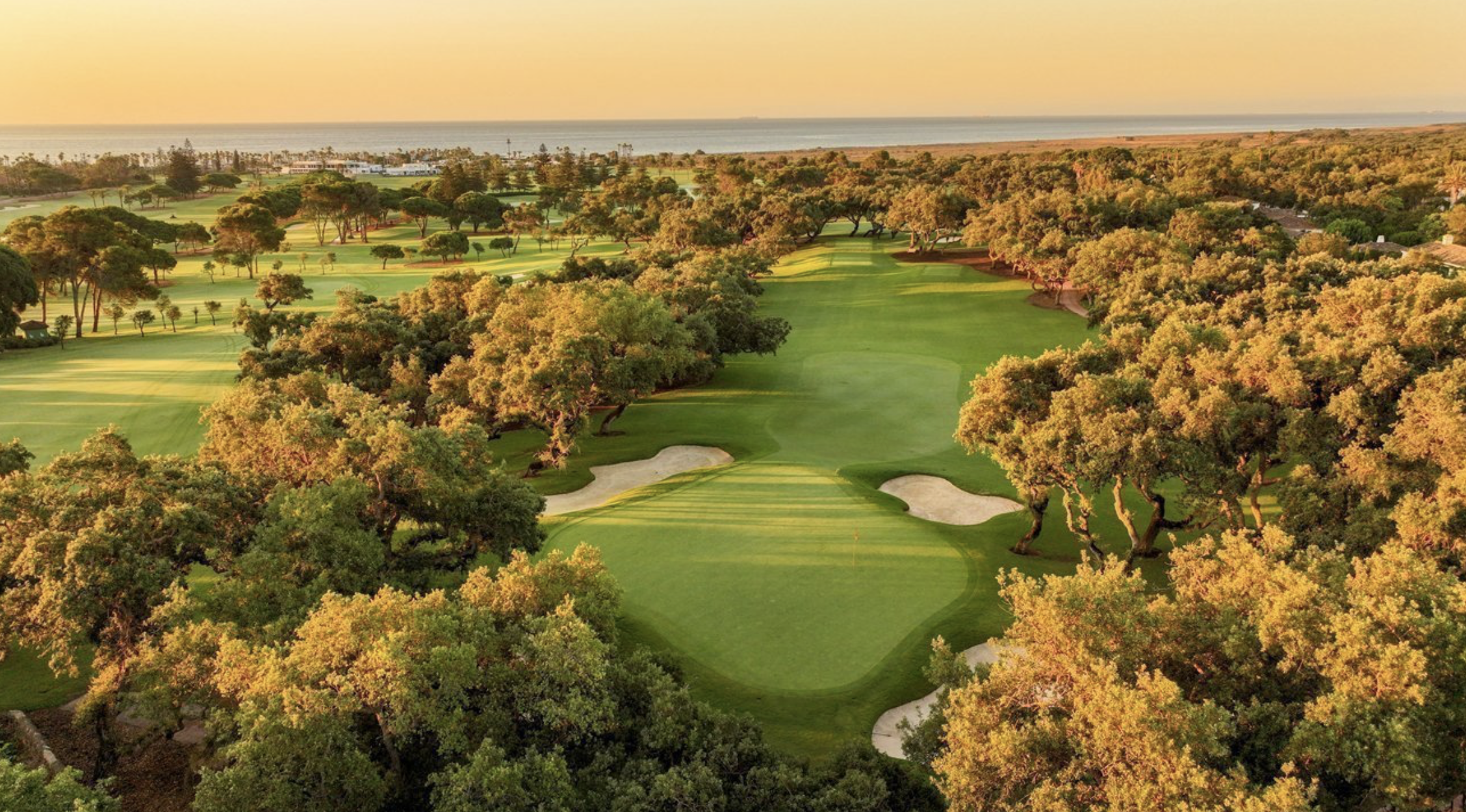 Sunset over a scenic golf course with lush green fairways, sand bunkers, and trees, with the ocean and horizon in the background. Caddie & Co. | Discover exclusive golf trips.