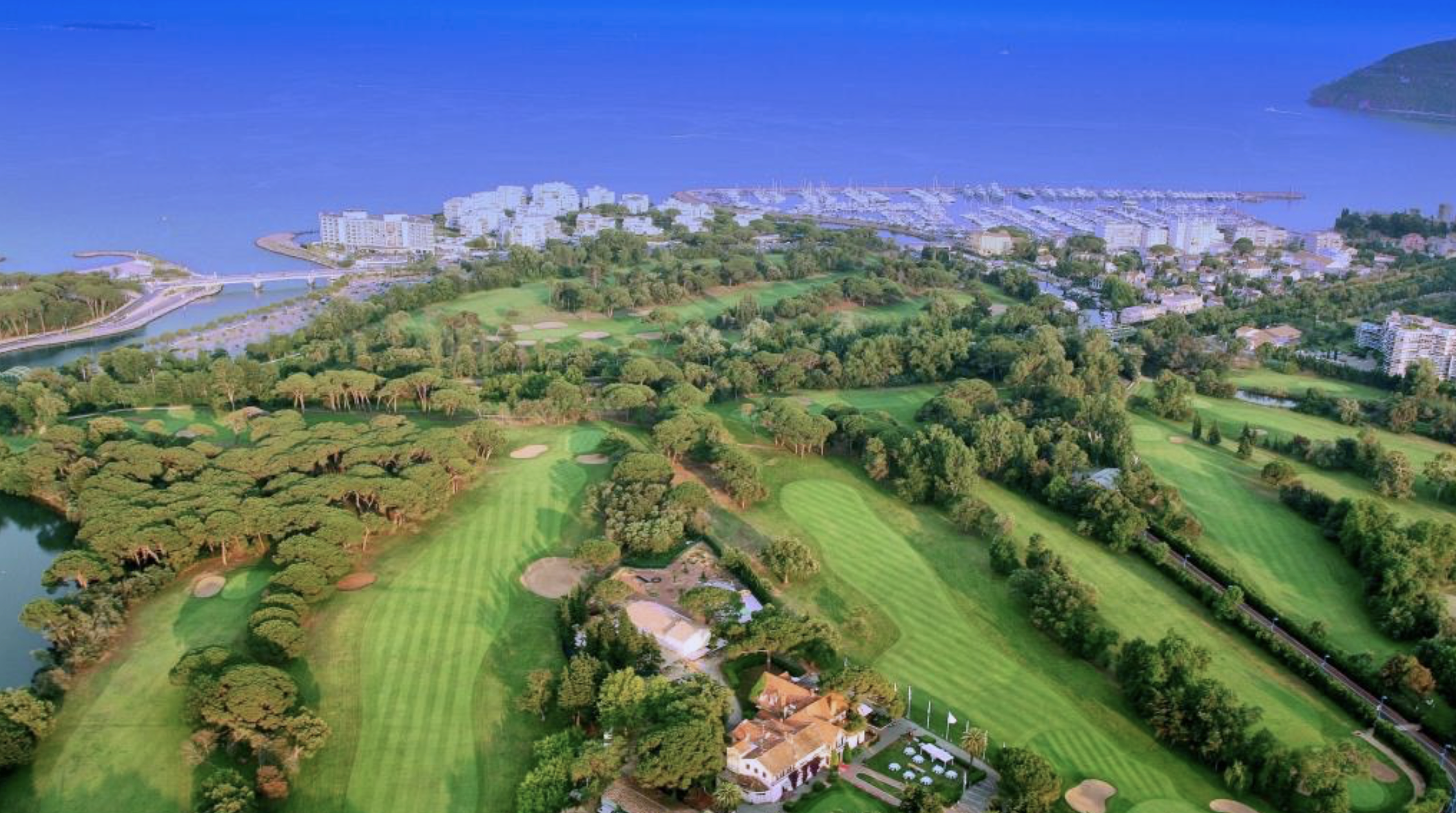 Aerial view of a golf course near a body of water with a marina and high-rise buildings in the background, surrounded by green landscape and trees.