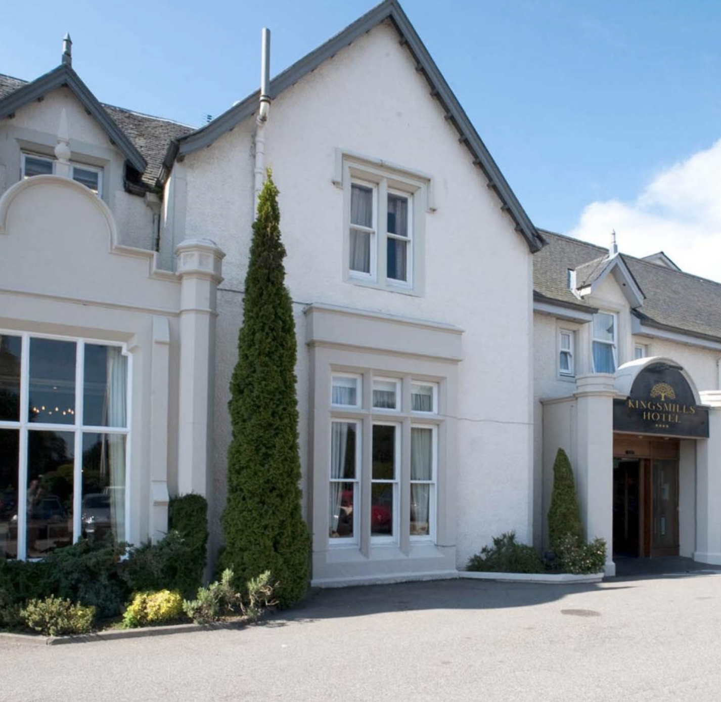 Exterior of a white hotel building with large windows, tall green trees, and a black and gold sign reading 'Kings Mills Hotel' on a bright, sunny day. Caddie & Co. | Discover exclusive golf trips.