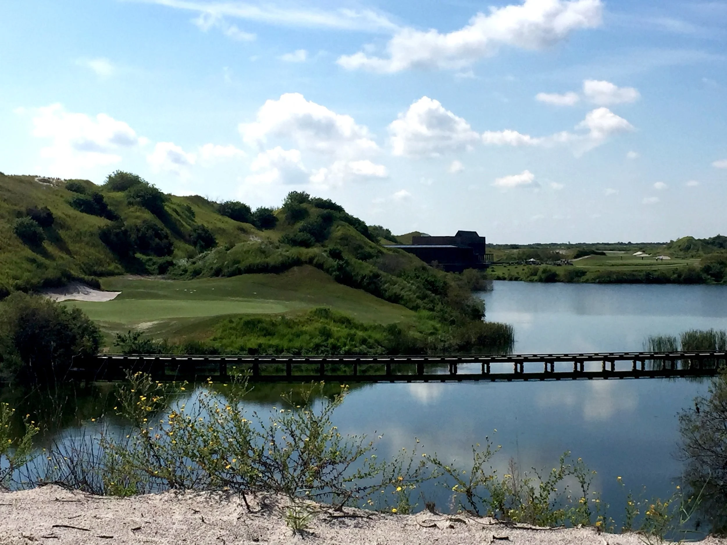 Scenic view of a golf course near a water body, with green hills, a bridge over water, and a building in the distance under a partly cloudy sky.