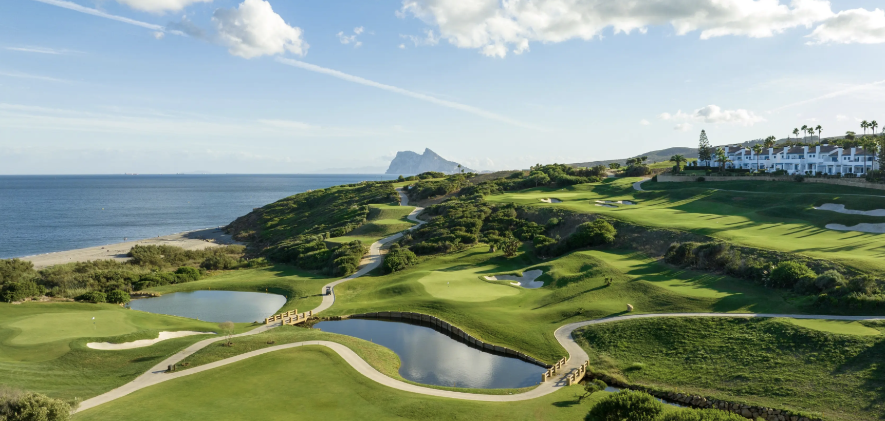 A scenic view of a golf course with green fairways, bunkers, water hazards, and walking paths, overlooking the ocean and distant mountains on a sunny day. Caddie & Co. | Discover exclusive golf trips.