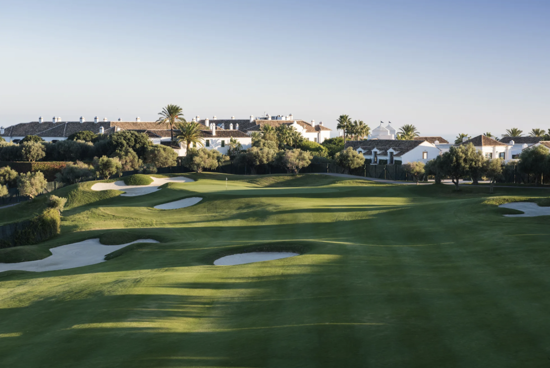 A golf course with sand bunkers, green grass, trees, and residential houses with white walls and brown roofs in the background under a clear sky.