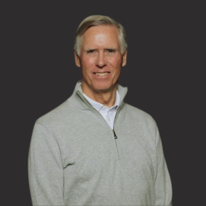 A smiling middle-aged man with gray hair wearing a light gray quarter-zip sweater over a collared white shirt, standing against a dark background.
