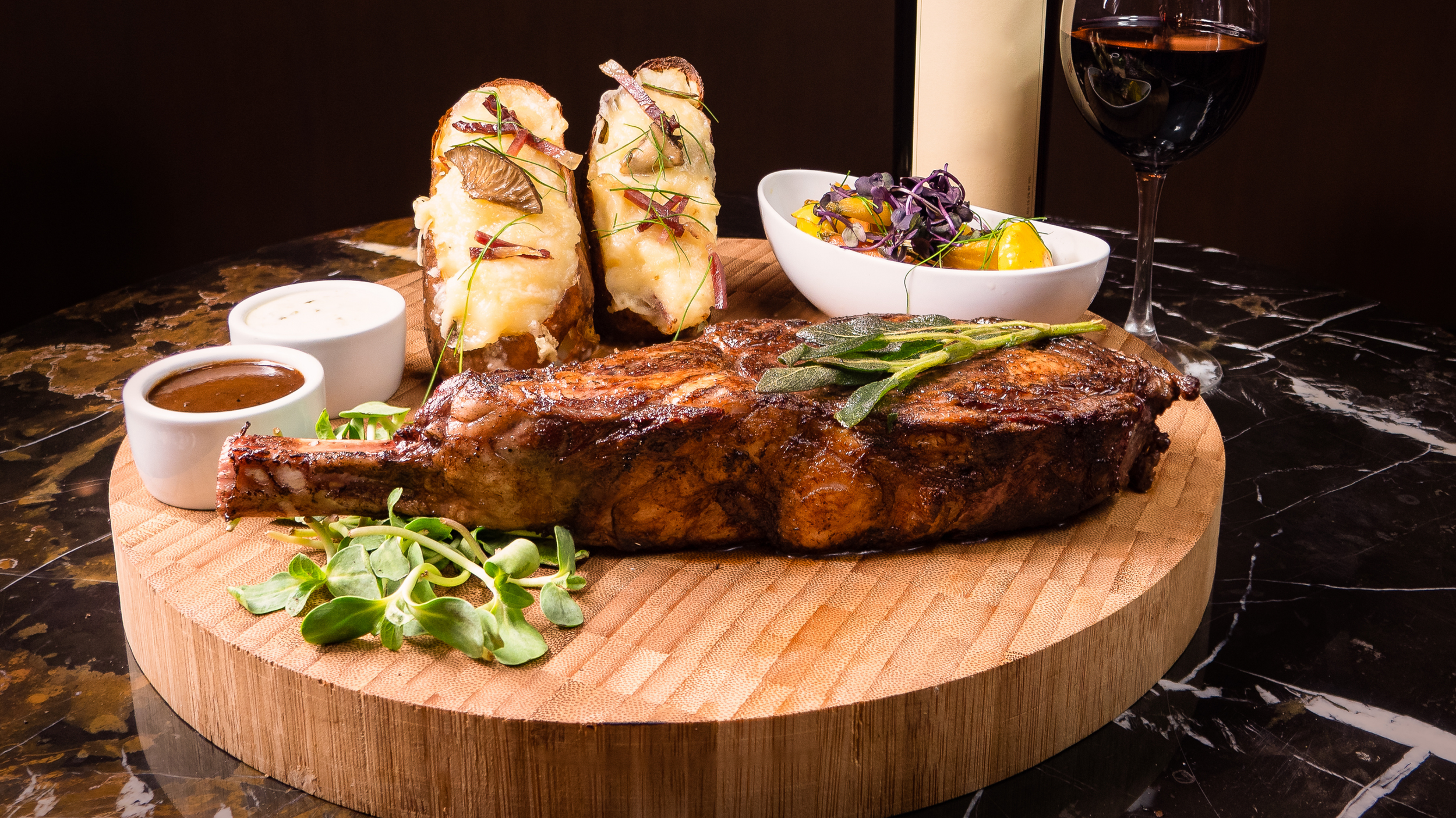 A roasted rack of lamb garnished with herbs on a wooden serving board, accompanied by mashed potatoes, three dipping sauces, a small salad in a white bowl, and a glass of red wine.