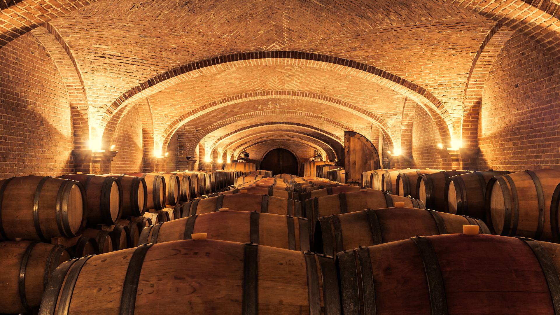 Wine cellar with brick vaulted ceiling and wooden barrels stacked in rows.
